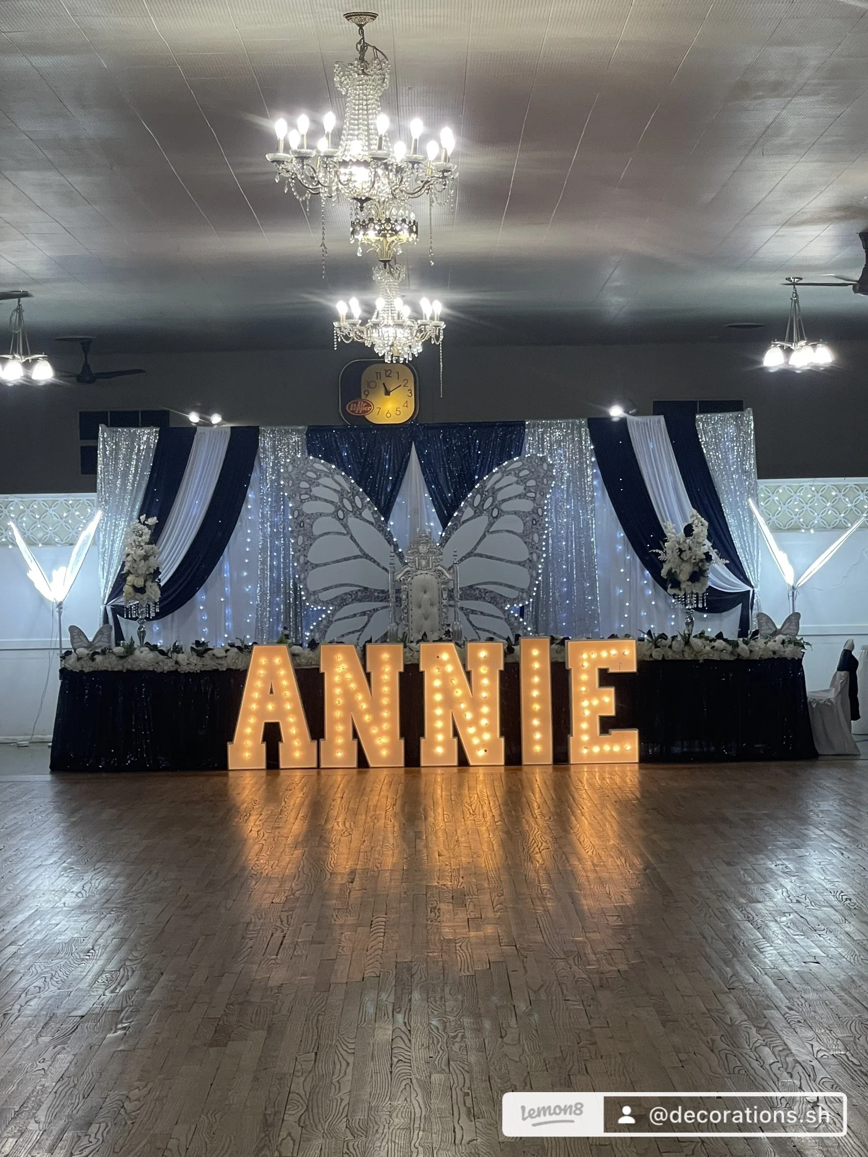 Wedding or event reception decorated with a large illuminated sign spelling 'ANNIE' in front of a black and white butterfly-themed backdrop, featuring flowers, drapes, and fairy lights, with chandeliers hanging from the ceiling.
