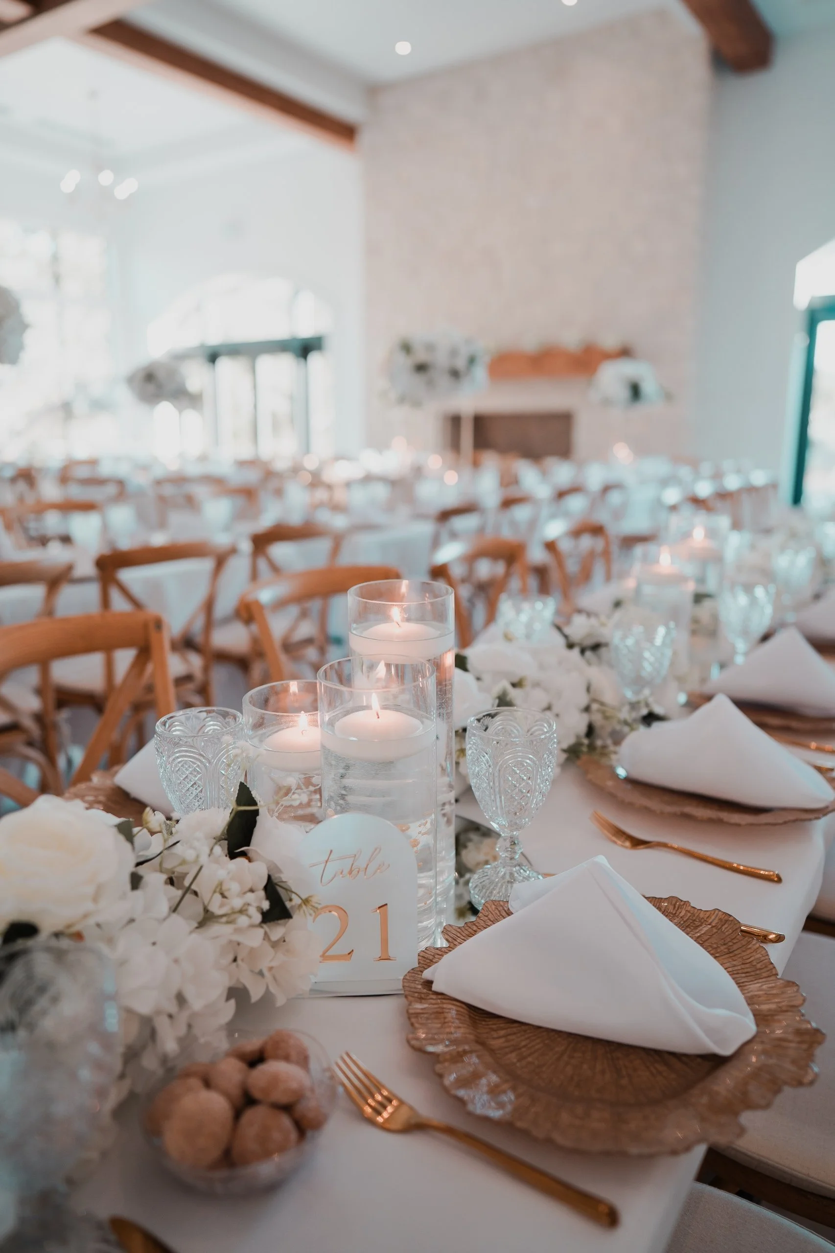 Elegant wedding reception table with white floral arrangements, candles in glass holders, gold utensils, and white napkins on textured plates, with a rustic interior and large windows in the background.