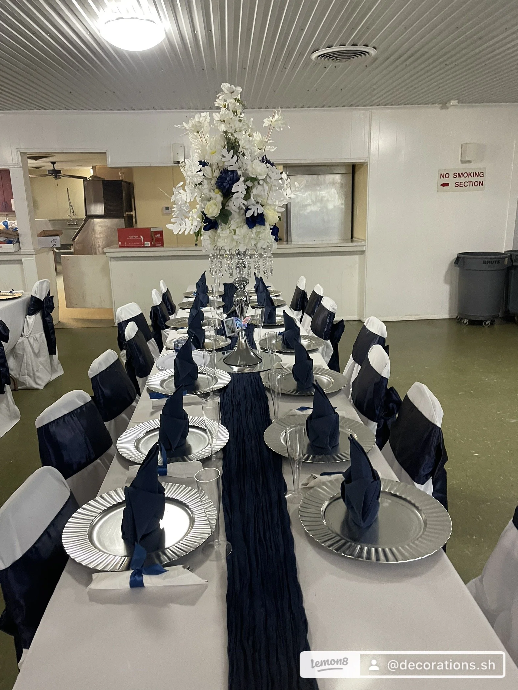 A banquet table decorated for an event with navy blue and white accents, featuring a tall floral centerpiece with white and dark blue flowers, crystal adornments, navy blue napkins folded into standing shapes, white and navy chair covers, and place s