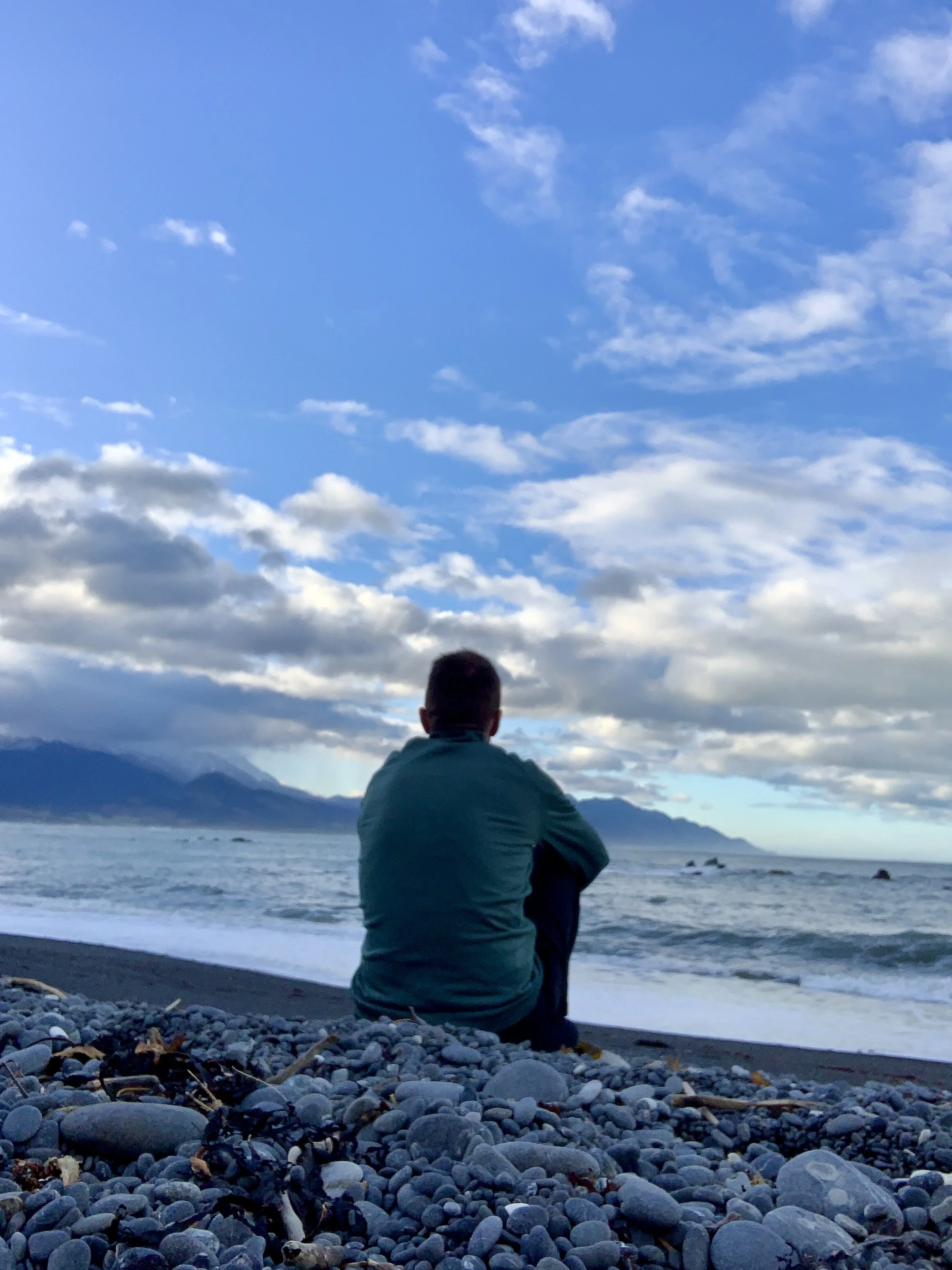 A person sitting on a pebble beach facing the ocean, with mountains in the distance and a partly cloudy sky above.