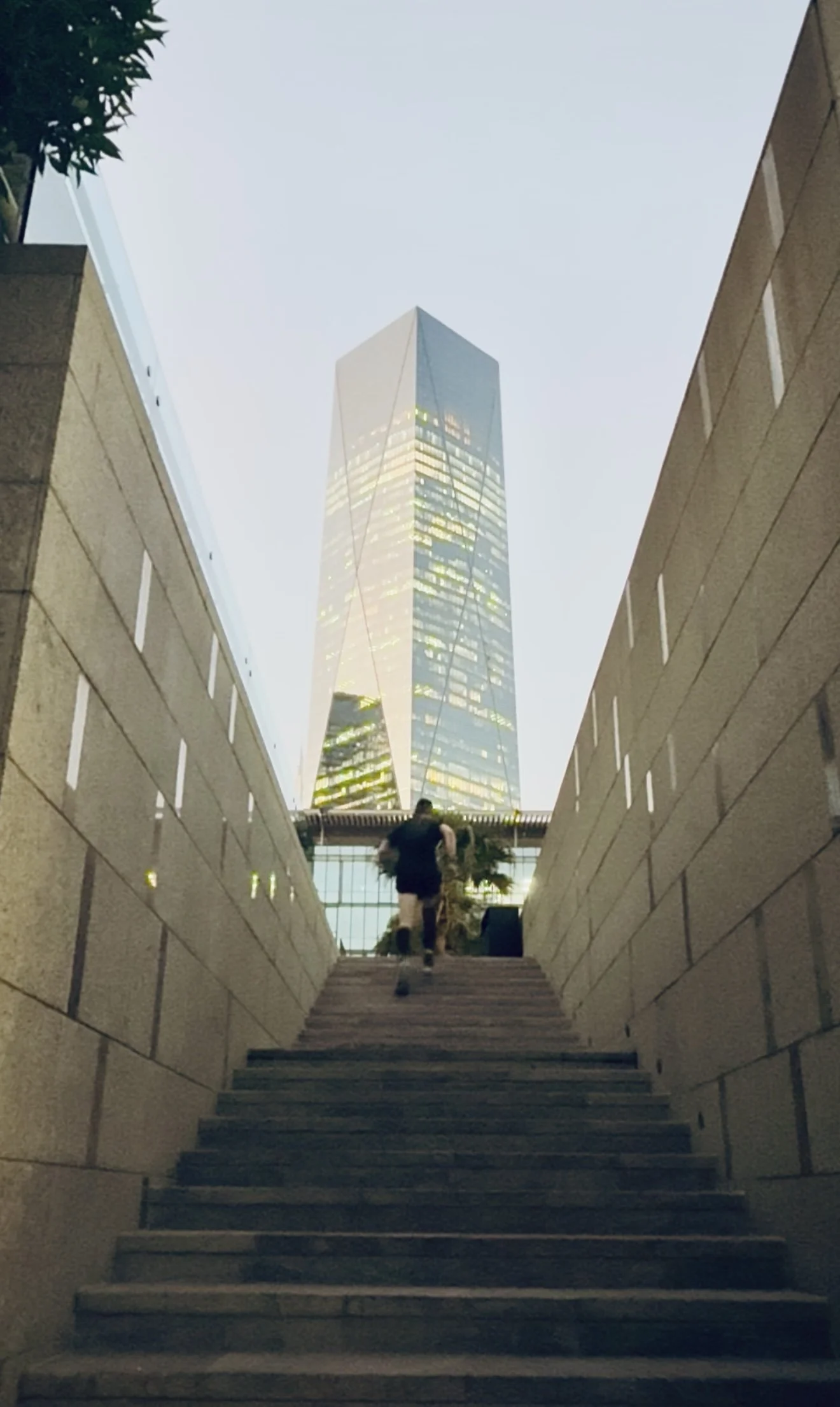 People climbing stairs between two brick walls towards a glass skyscraper in a city at dusk.