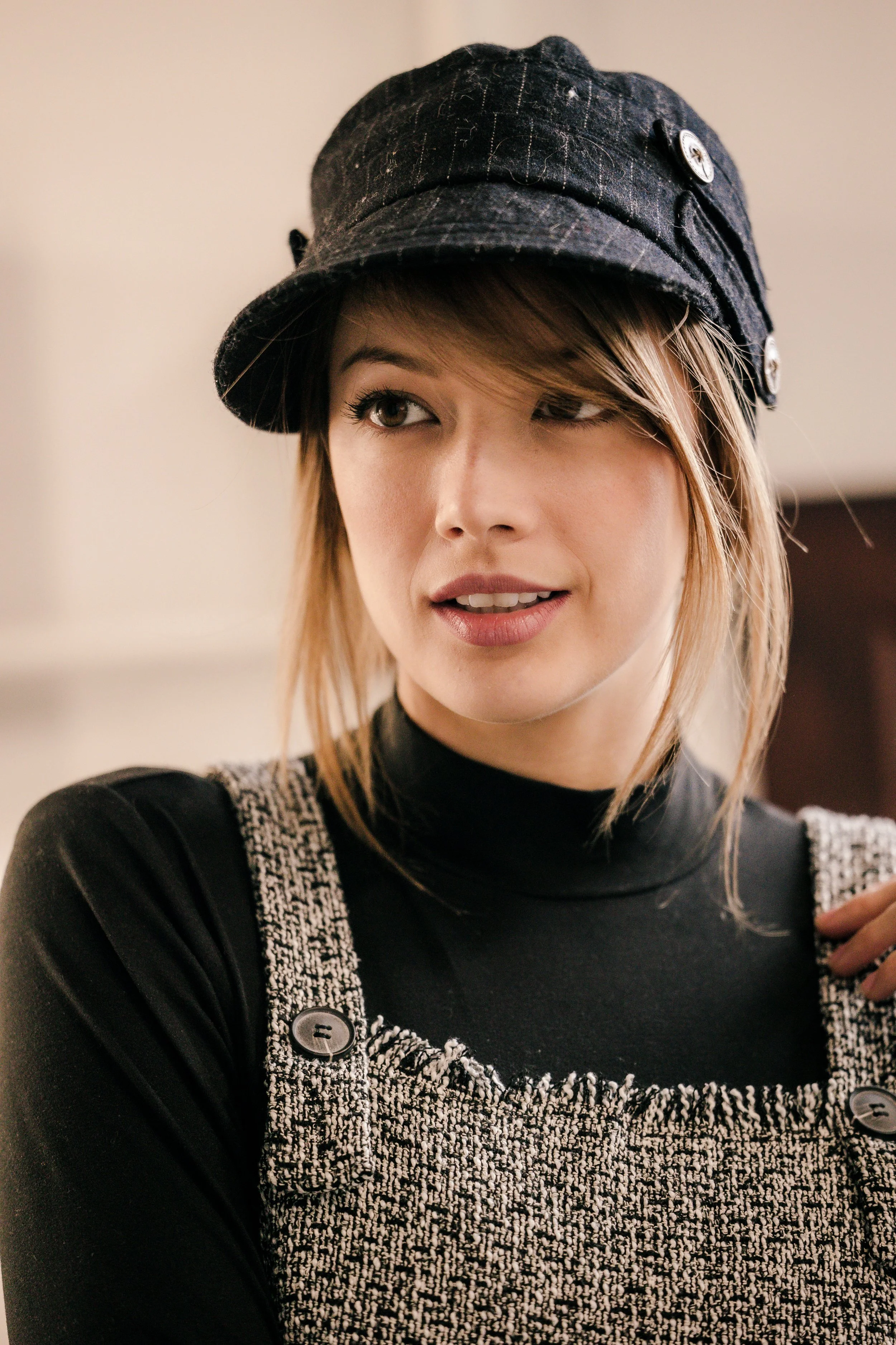 Young woman with light brown hair wearing a black hat with buttons, a black turtleneck, and a textured, black and white patterned overall, looking to the side.