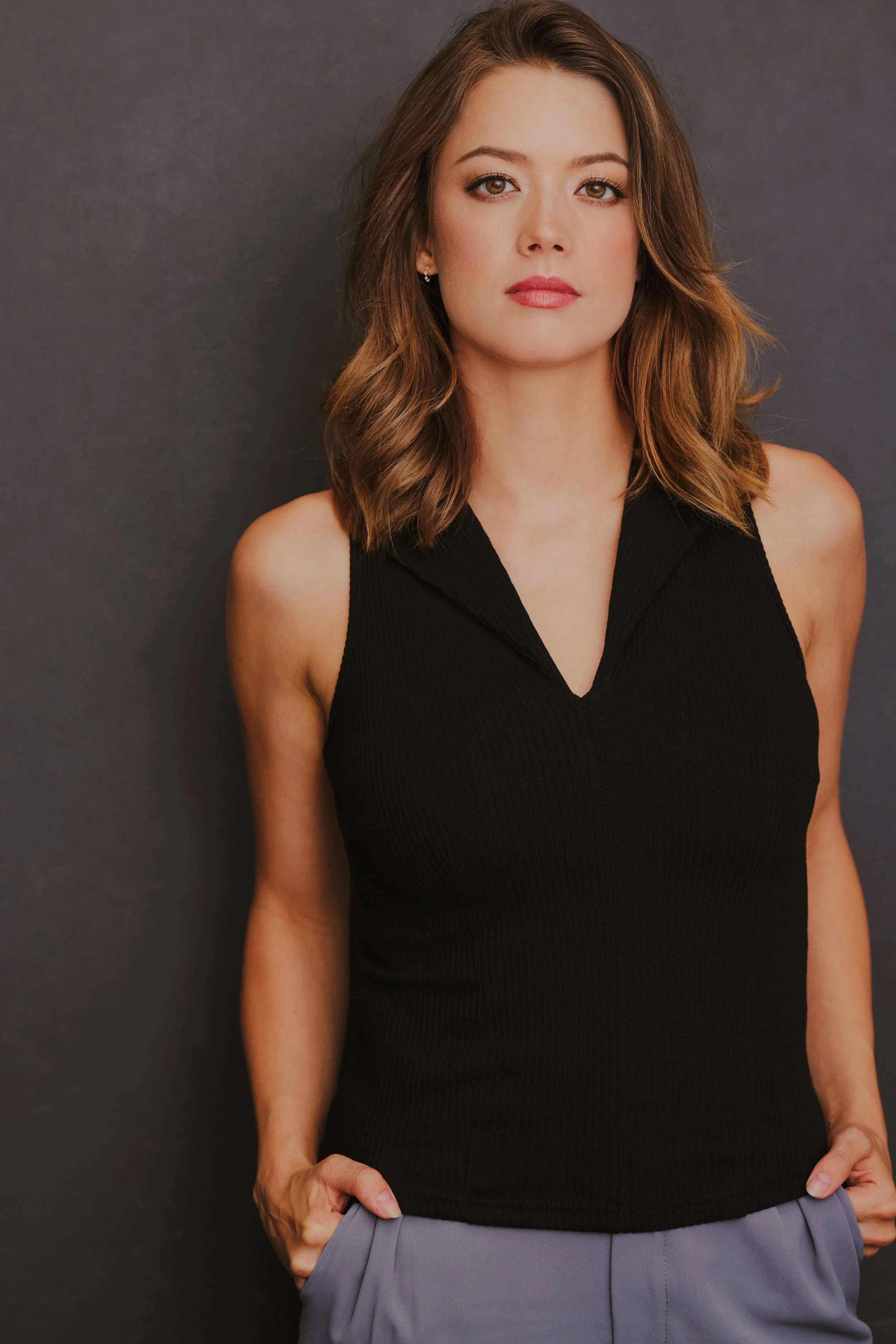 A woman with shoulder-length brown hair wearing a sleeveless black top and gray pants, standing against a gray background.