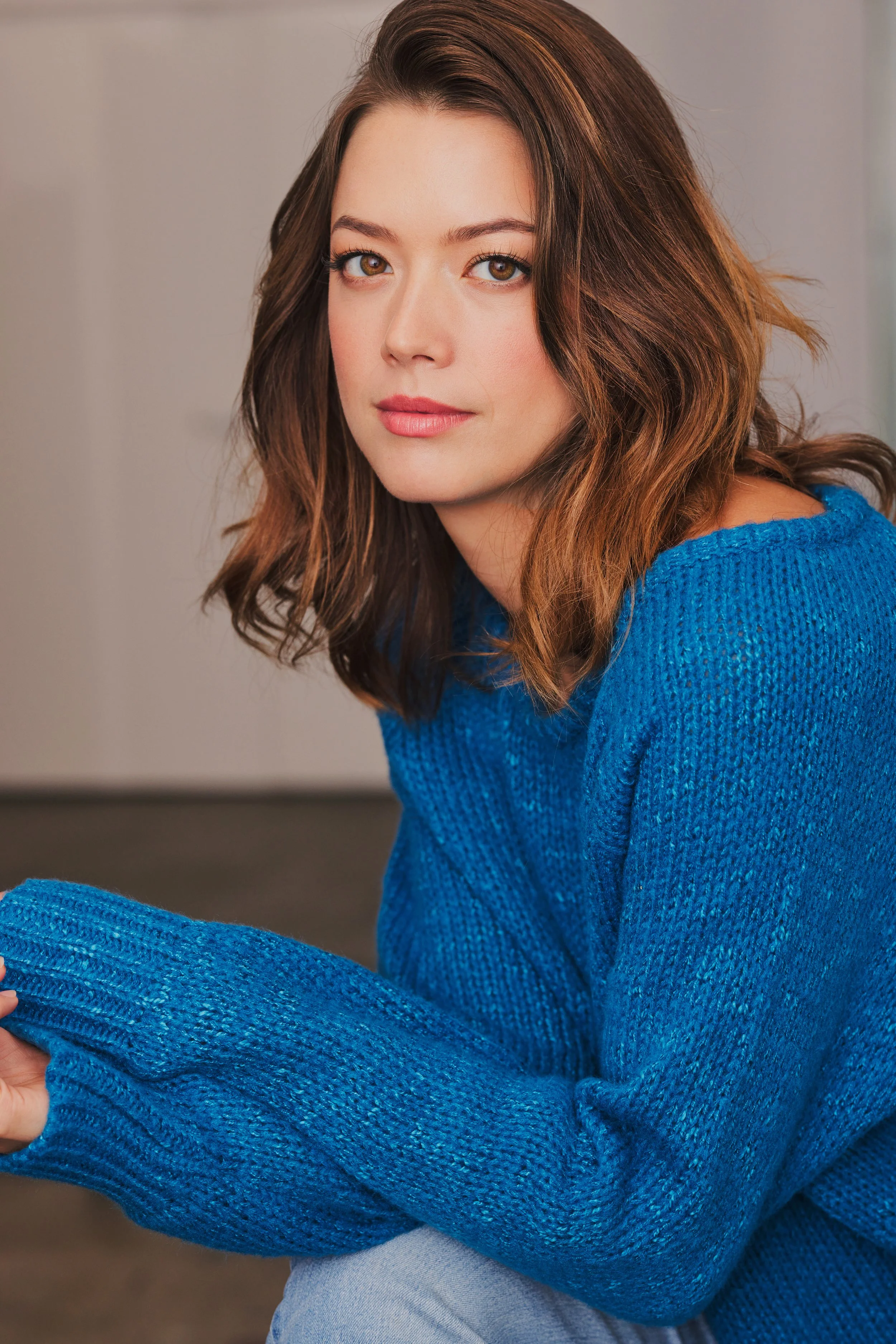 A young woman with shoulder-length brown hair wearing a blue knit sweater, sitting indoors with her arms crossed and looking at the camera.