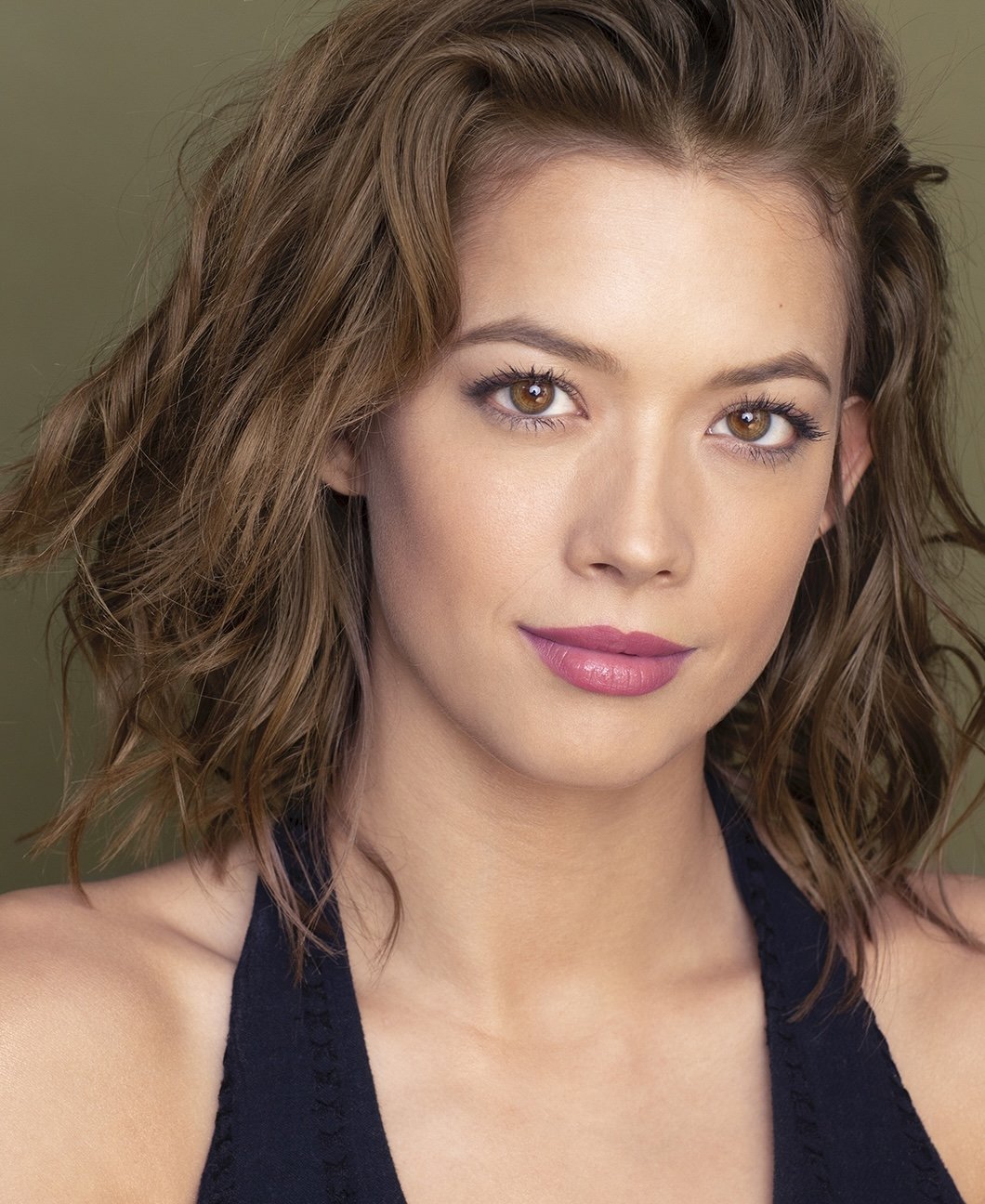 Close-up portrait of a young woman with wavy brown hair and brown eyes, smiling softly, wearing a black tank top against a muted green background.