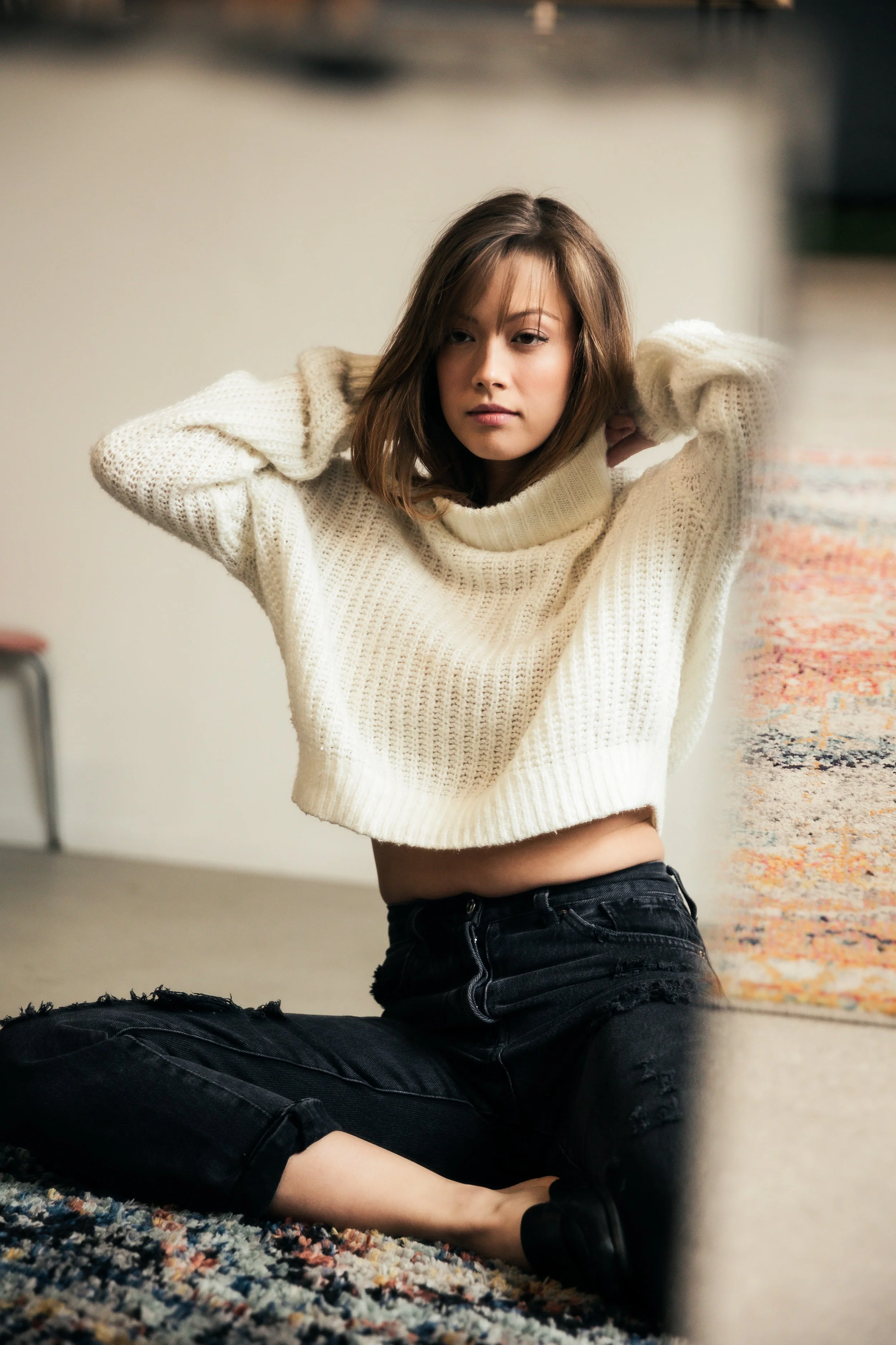 A young woman sitting on the floor, wearing a cream-colored knit sweater and distressed black jeans, with her hands behind her head and looking to the side.