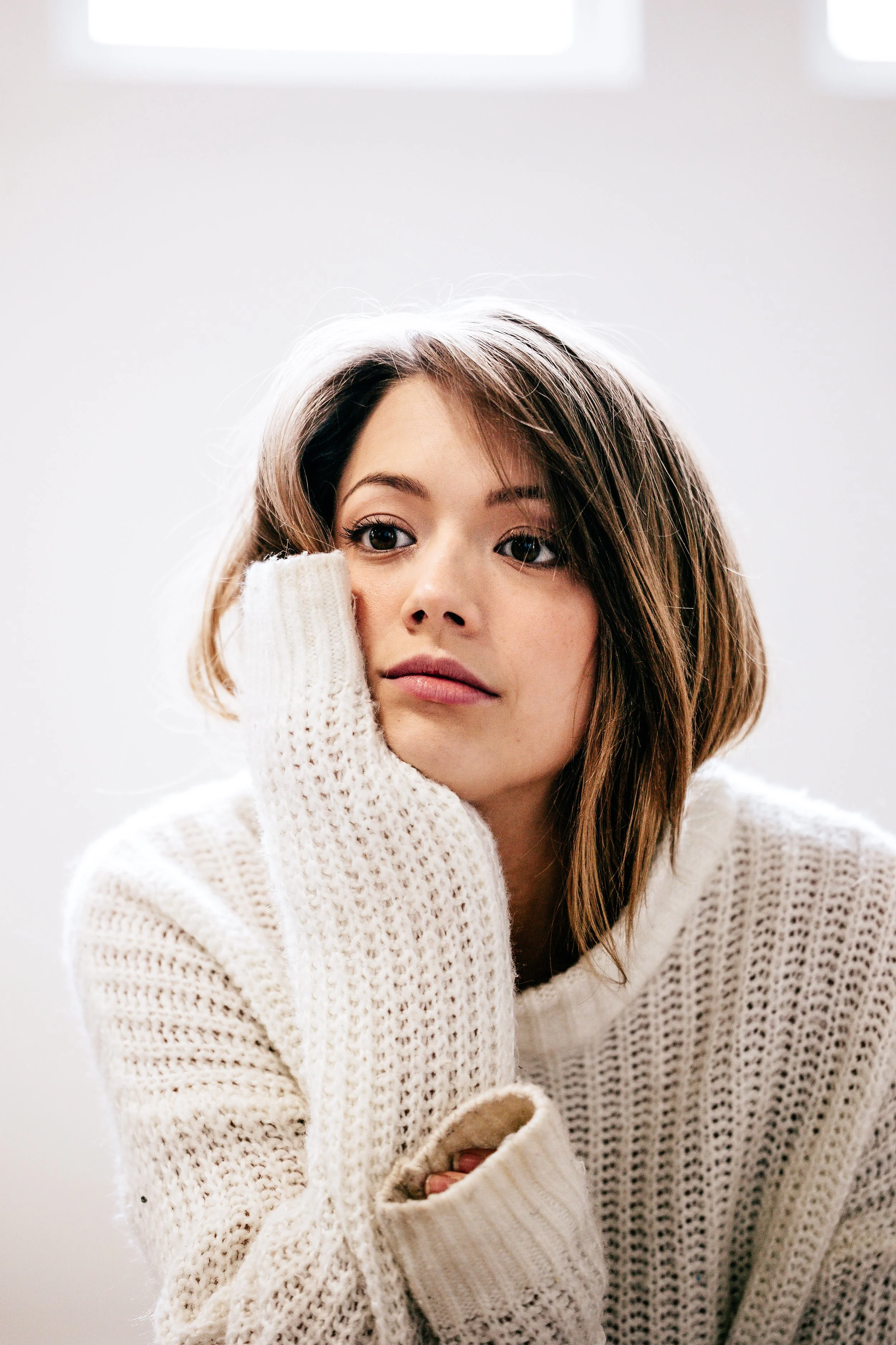 A young woman with short brown hair and light skin, resting her face on her hand, wearing a chunky white knit sweater, looking contemplative against a plain white background.