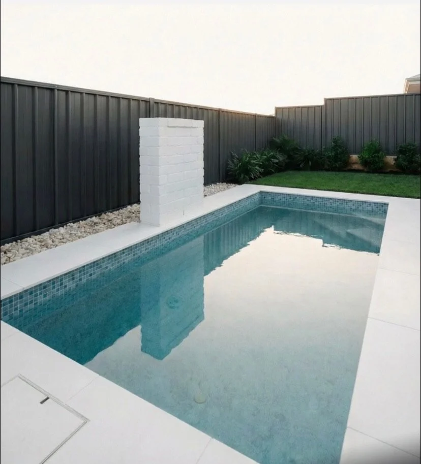 A small backyard swimming pool with a tall white brick structure at one end, surrounded by a black metal fence, green plants, and a well-manicured lawn.