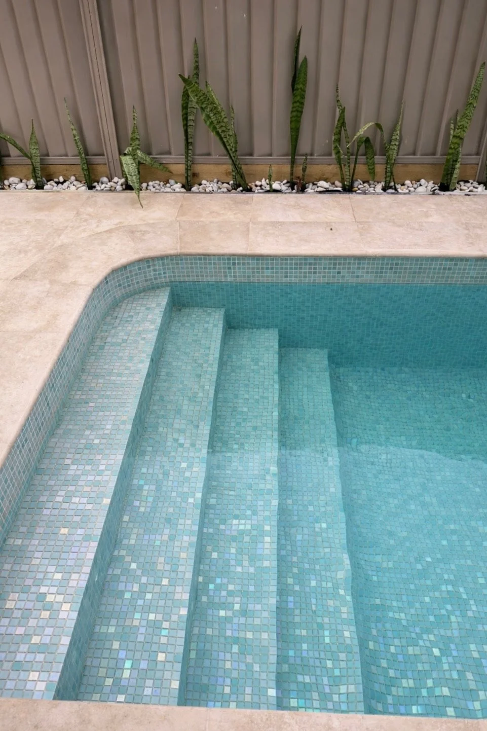 An indoor swimming pool with tiled steps leading into the water, surrounded by beige tiles and green plants along a fence.