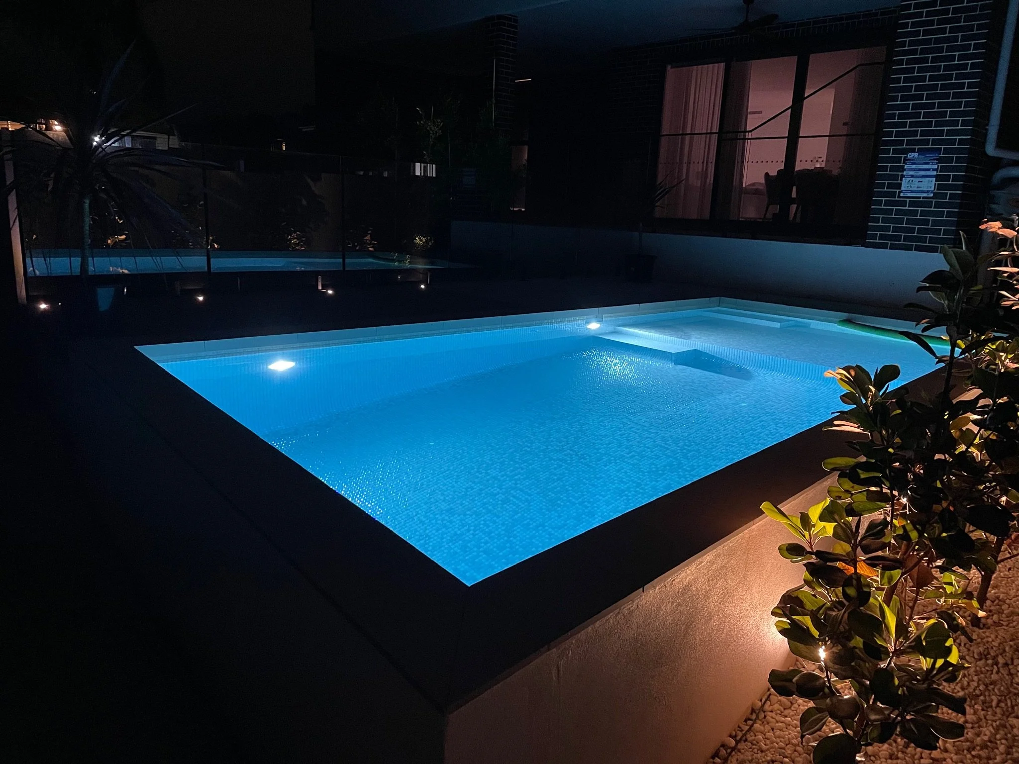 An illuminated swimming pool at night, with a clear, blue water surface and surrounding plants, adjacent to a building with a patio and a sliding glass door.