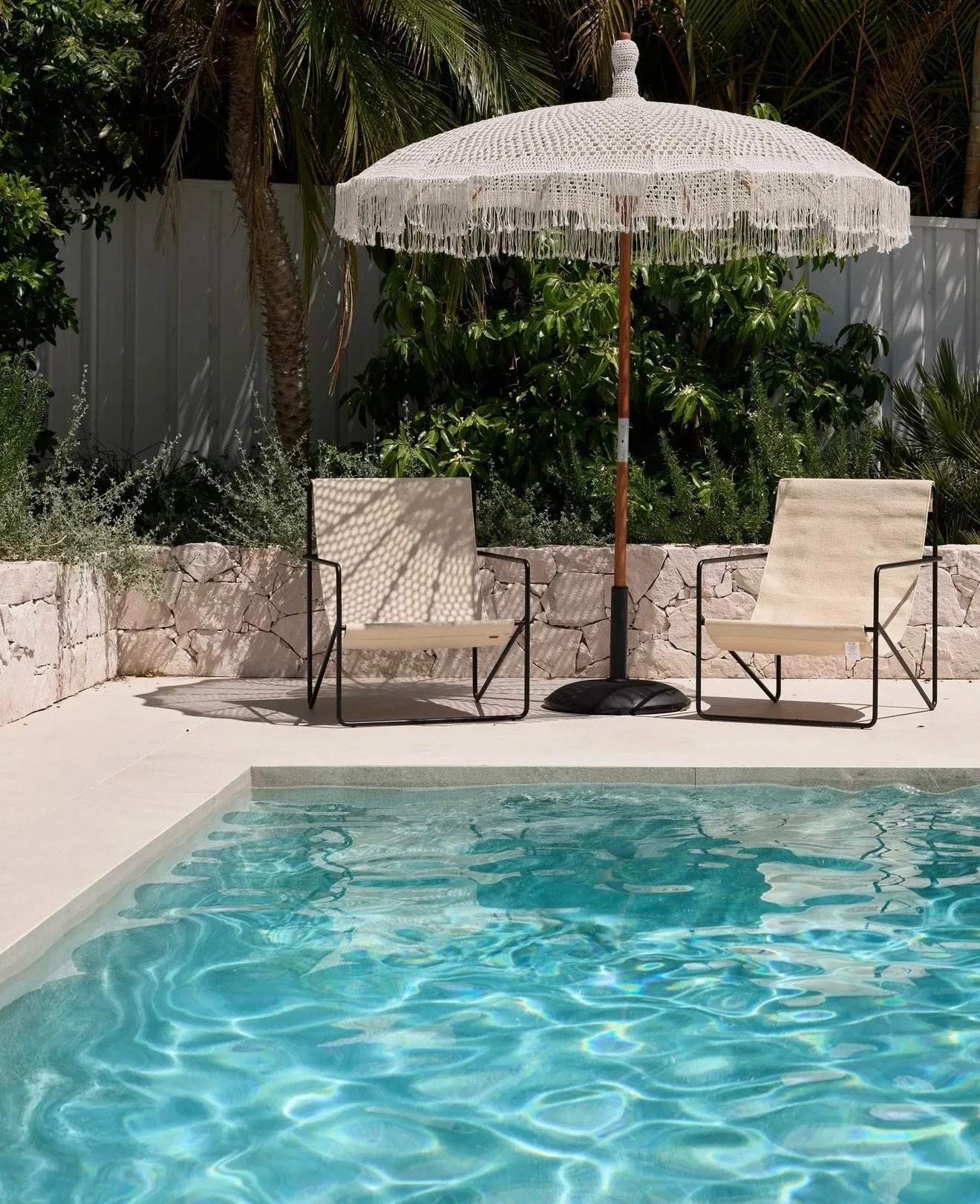 Swimming pool with two beige lounge chairs and a white umbrella on a pool deck, surrounded by green plants and a white fence.