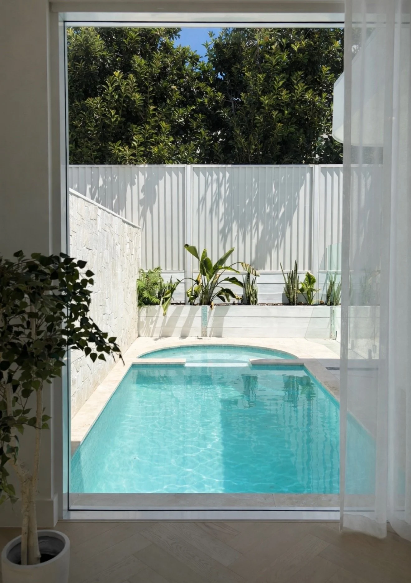 View of a small, rectangular swimming pool seen through a large sliding glass door, with a garden and tall green trees in the background. The pool area has a white fence and potted plants.
