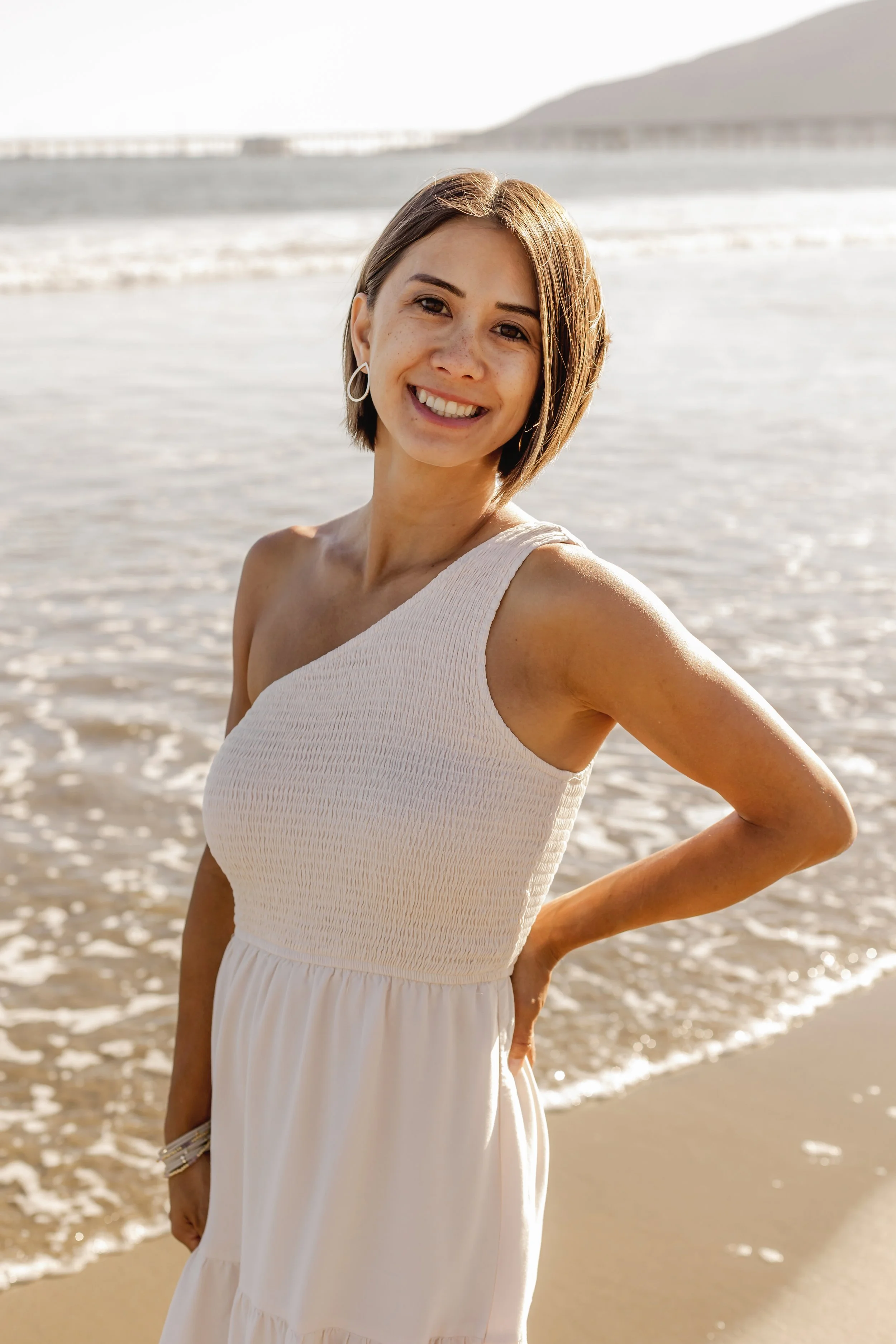 A woman with short brown hair is smiling and standing on a beach near the water, wearing a sleeveless white dress.