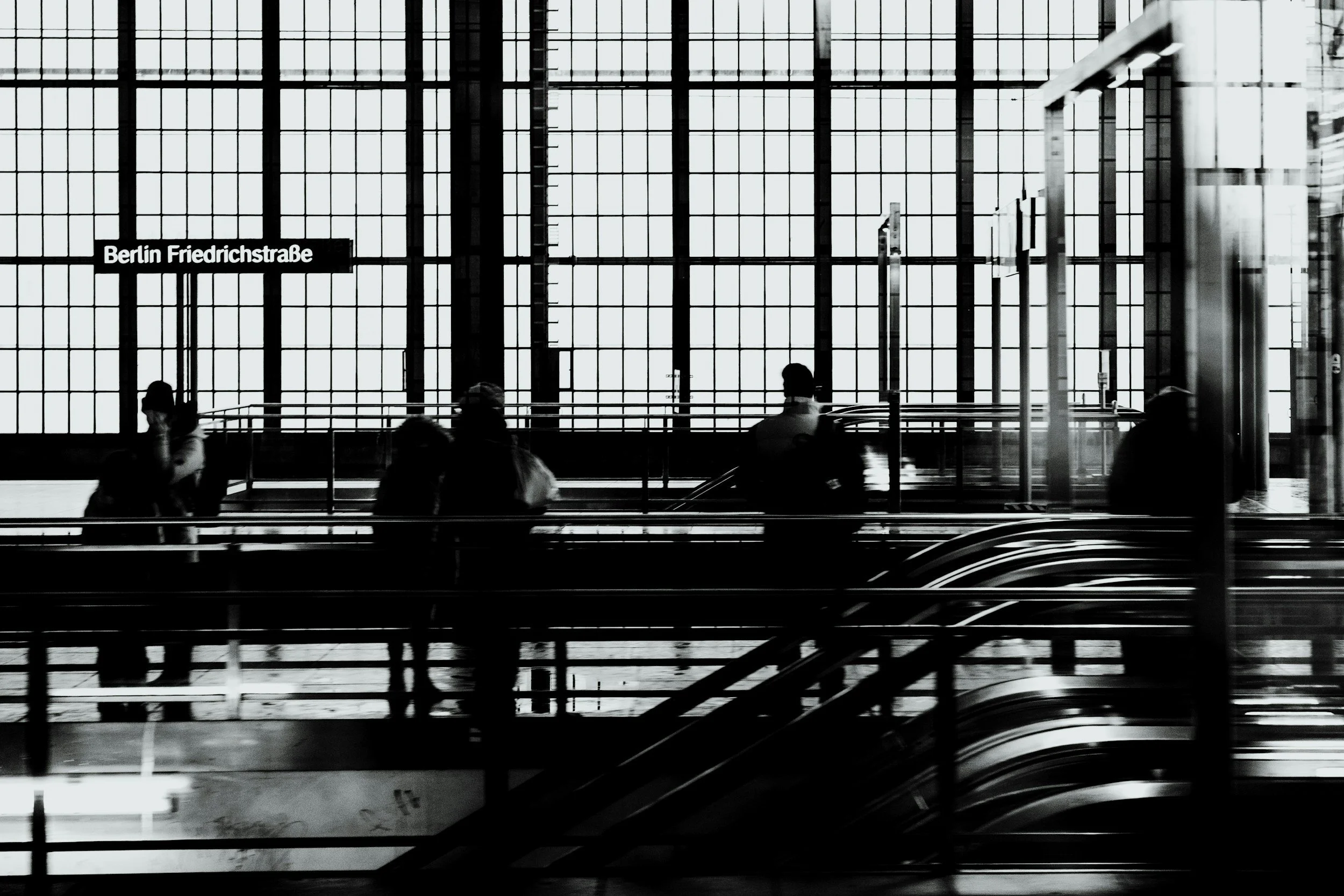 Black and white photo of interior of a modern building with large window panes, escalators, and four silhouettes of people sitting or standing, with a sign indicating Berlin Friedrichstraße.