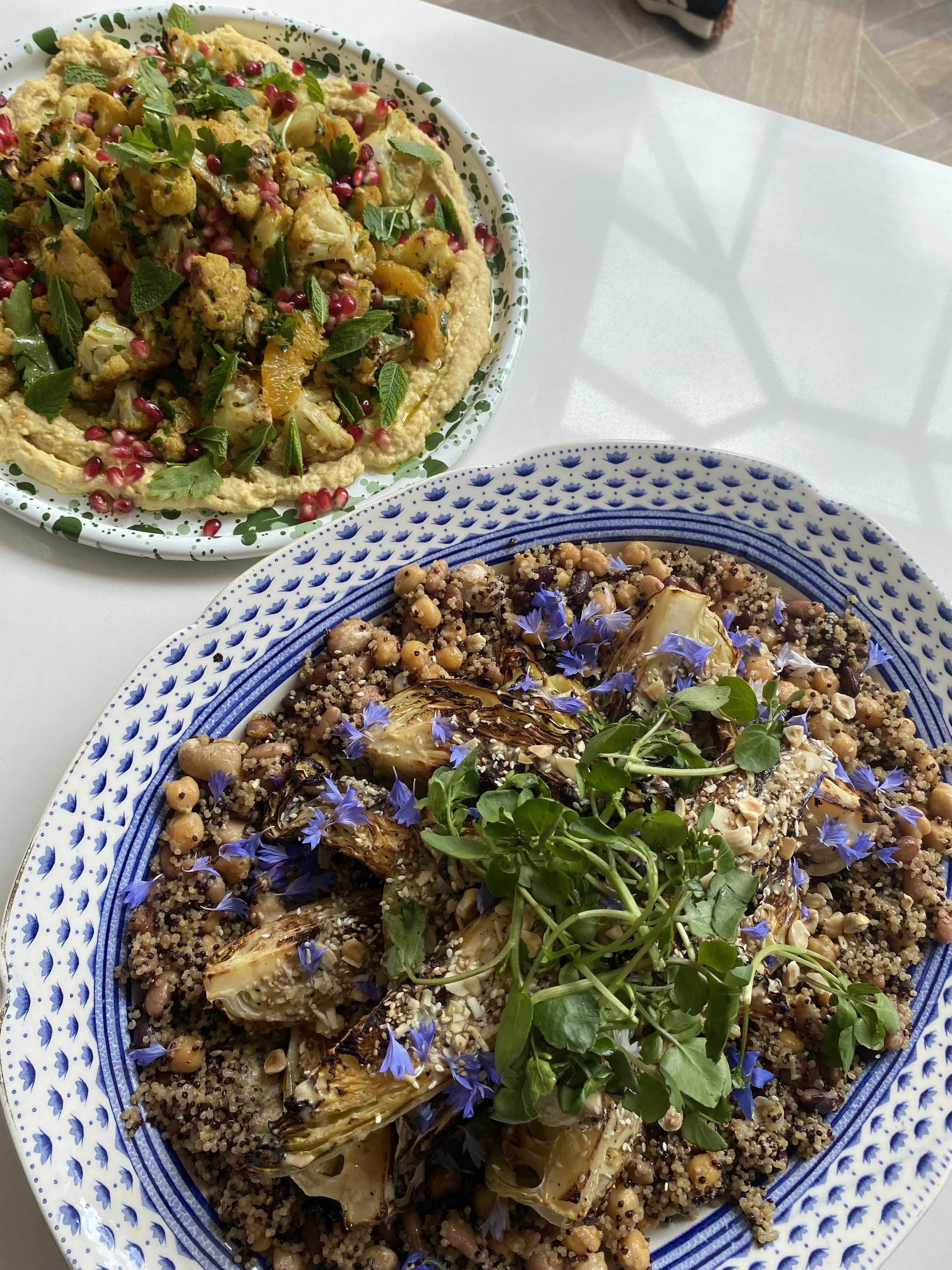Two plates of colorful vegetarian salad dishes on a white surface. Roasted vegetables, quinoa, chickpeas, with blue edible flowers and fresh greens. Creamy hummus garnished with herbs, pomegranate seeds, and spiced cauliflower.