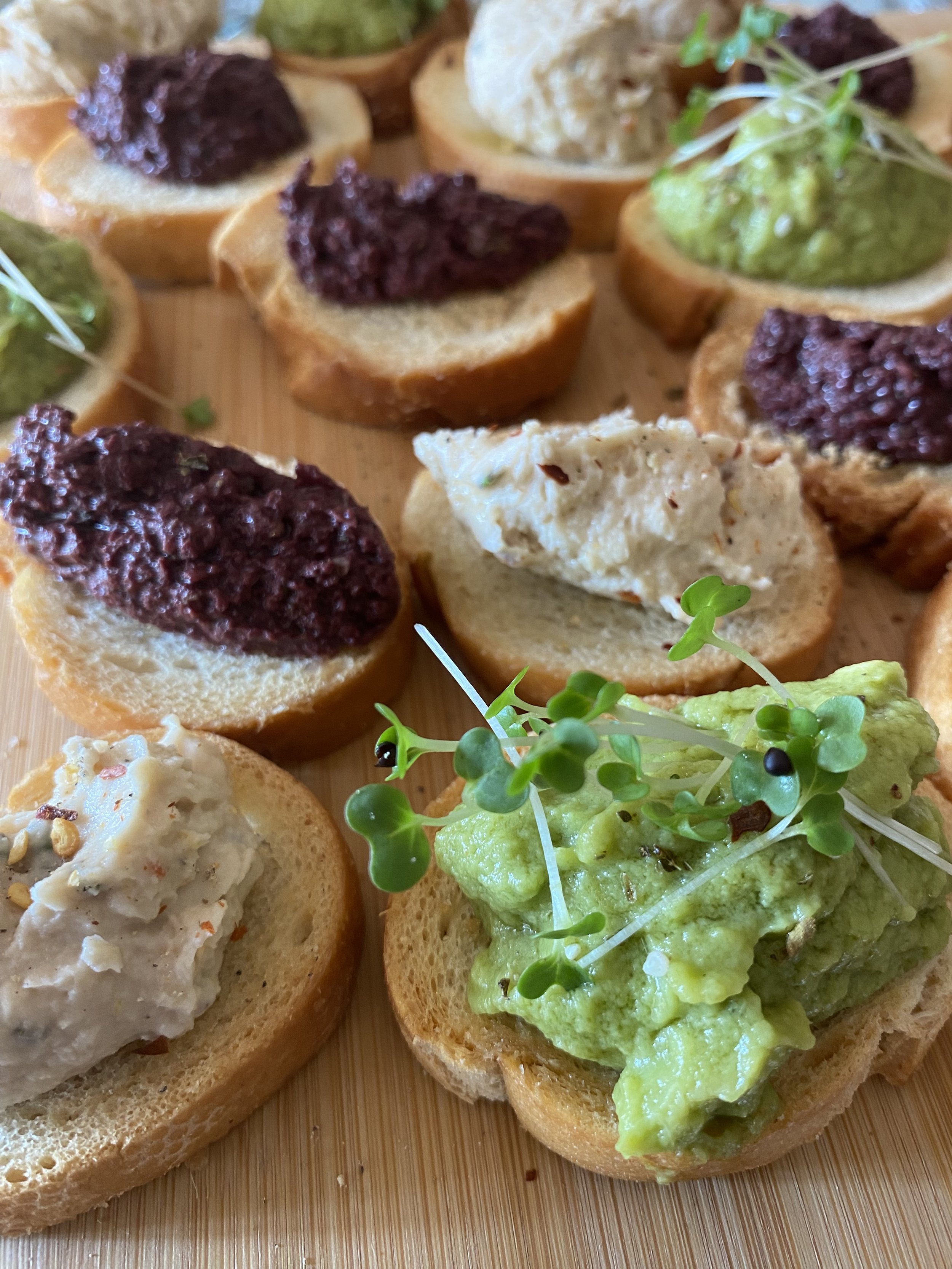 Close-up of assorted crostini with toppings such as tapenade, guacamole, cheese spread, and herbs on a wooden surface.
