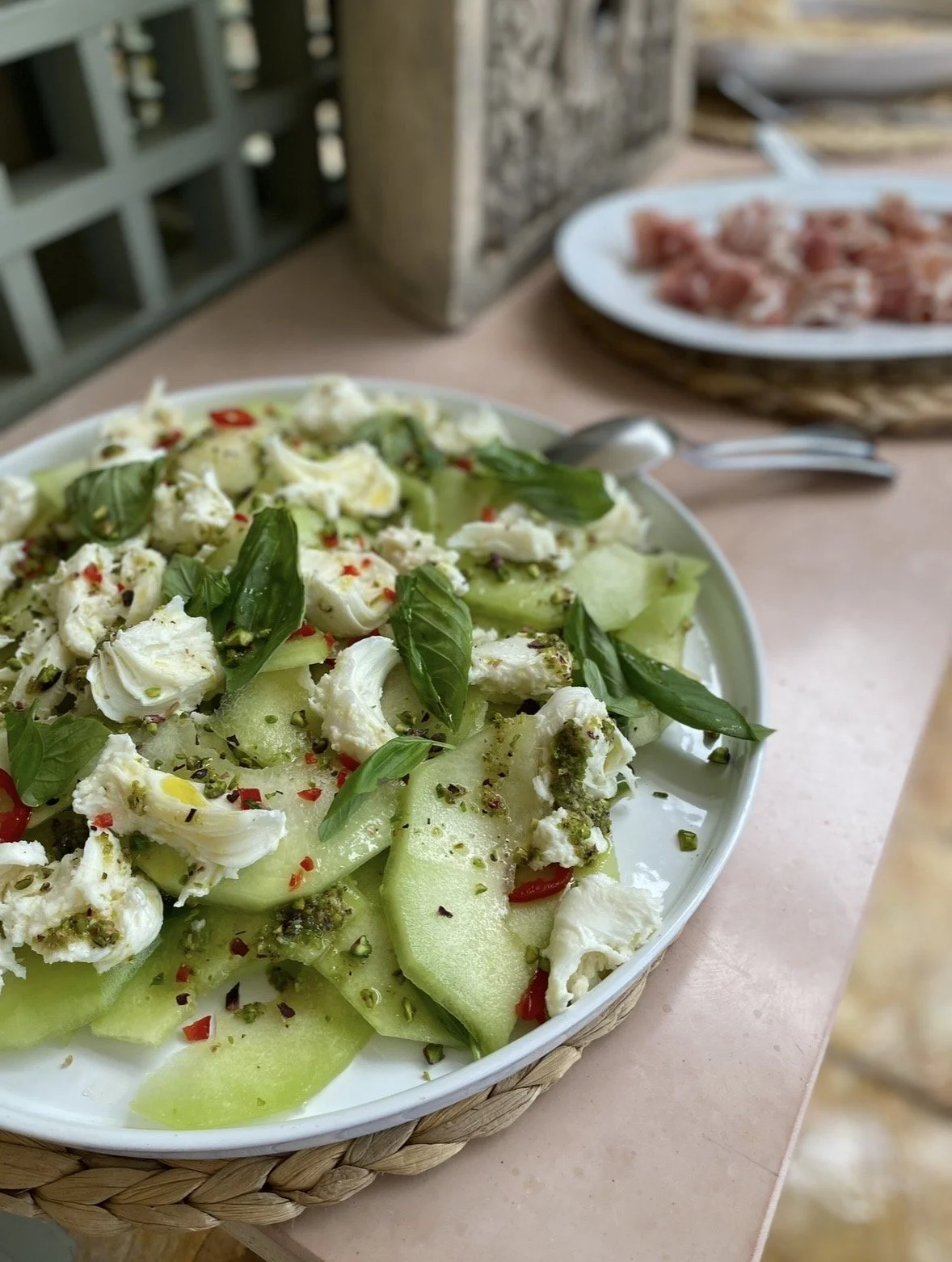 A plate of sliced honeydew melon and cucumber topped with torn mozzarella, basil leaves, red pepper flakes, and black pepper on a pink table.