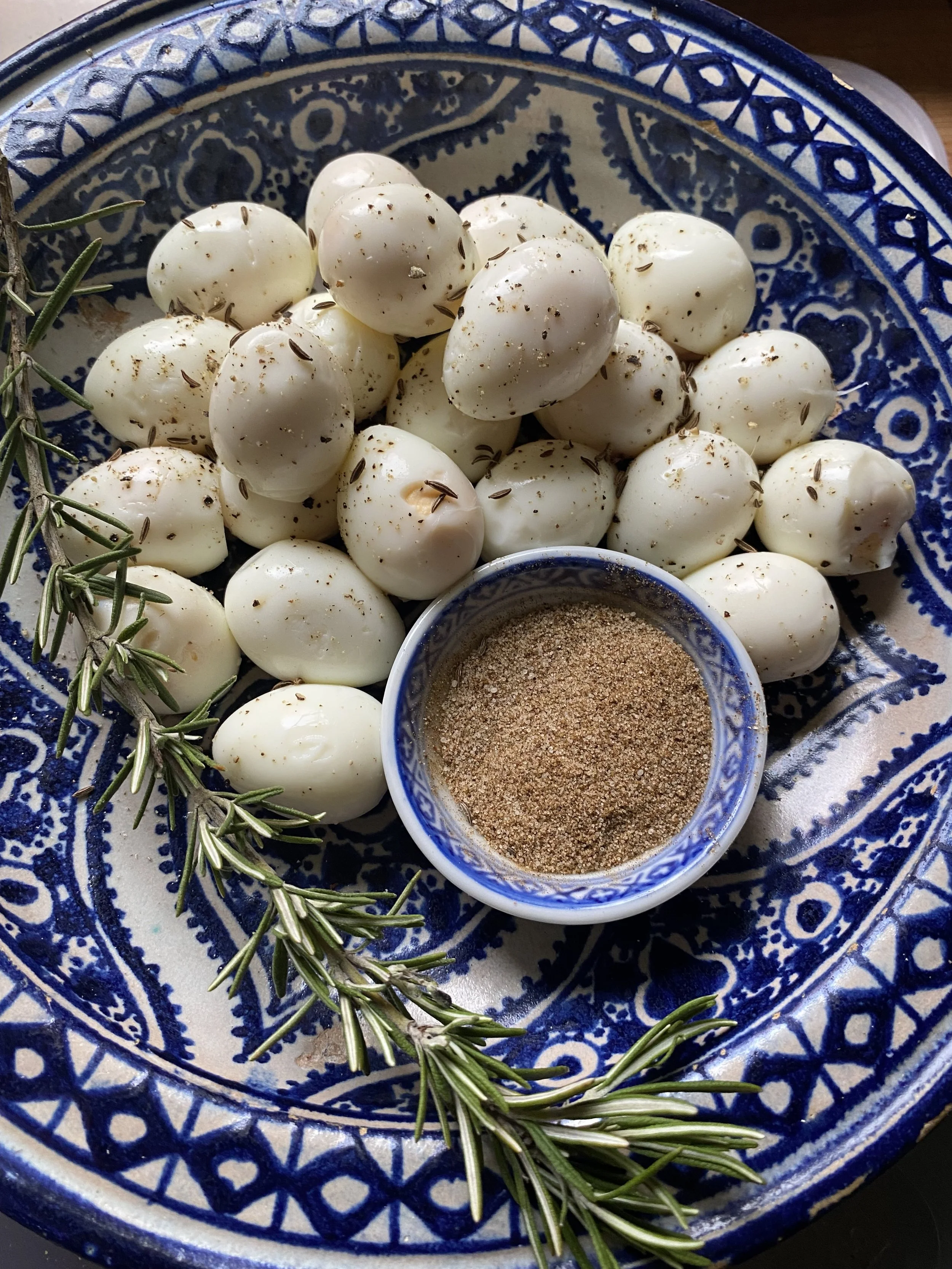 A decorative blue and white ceramic plate with peeled hard-boiled eggs sprinkled with herbs, a small bowl of black pepper, and sprigs of fresh rosemary.