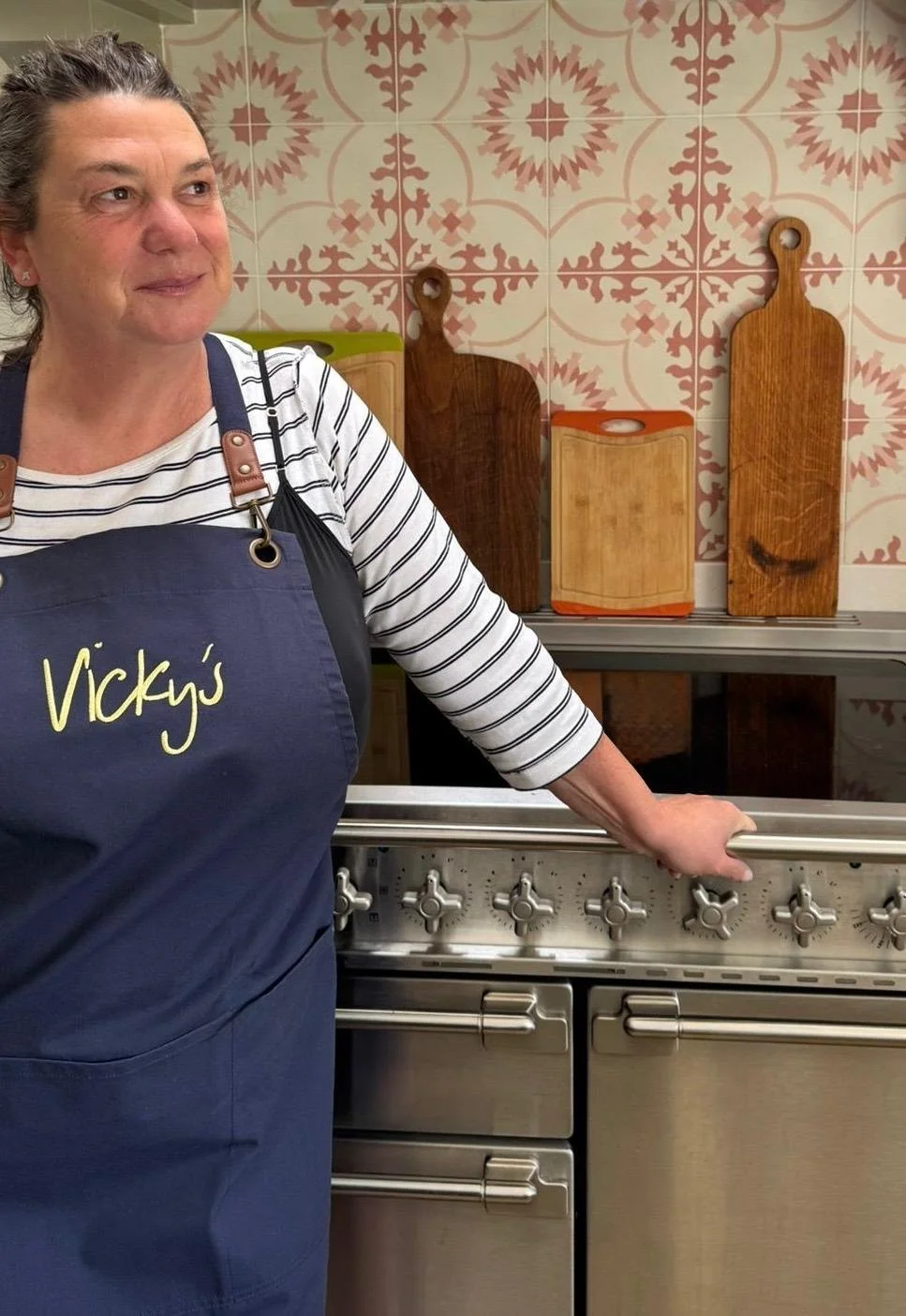 A woman standing in a kitchen, wearing a striped shirt and an apron with the words "Vicky's" embroidered on it. She has her hand resting on a stainless steel stove.