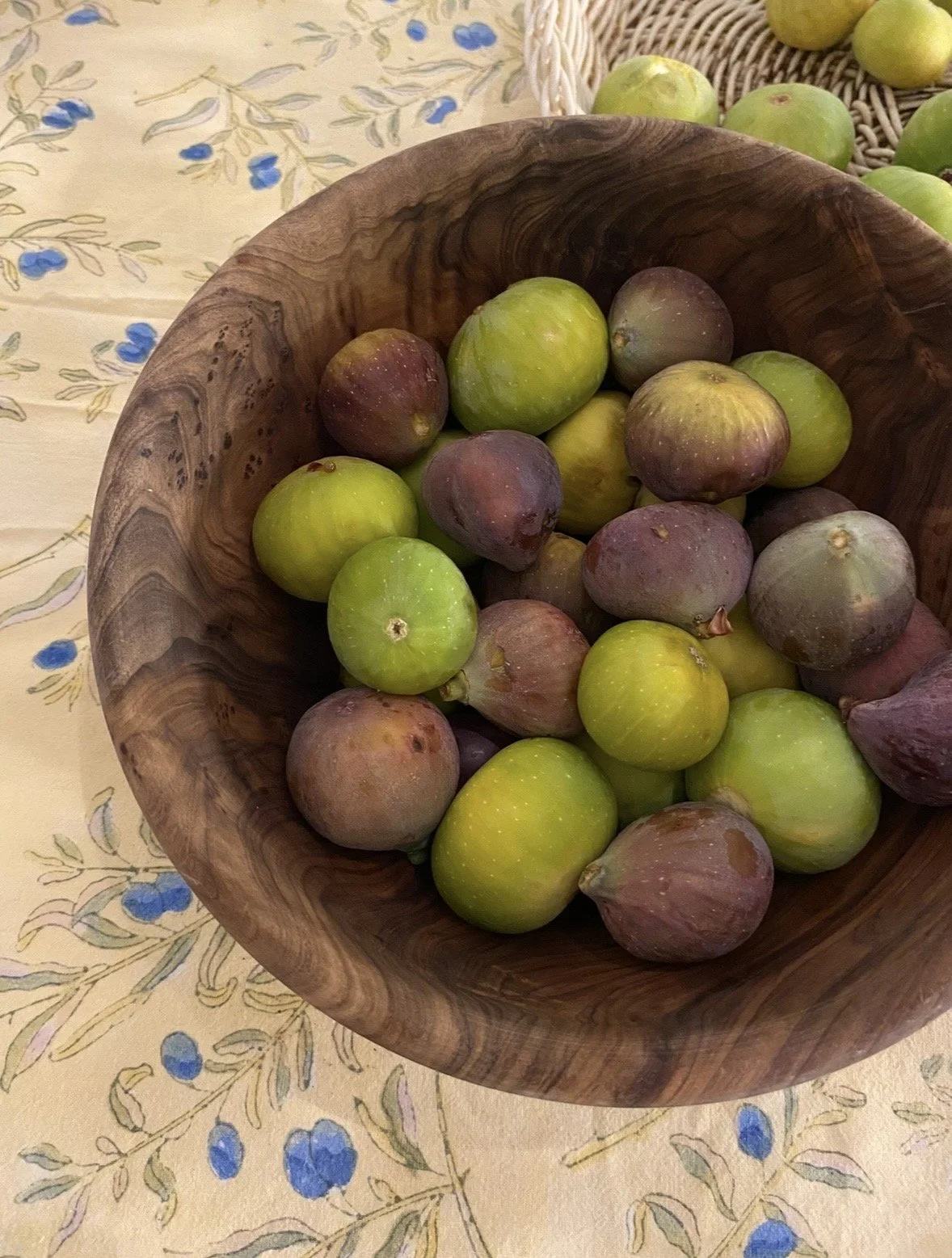 A wooden bowl filled with green and purple figs sits on a table with a floral tablecloth.