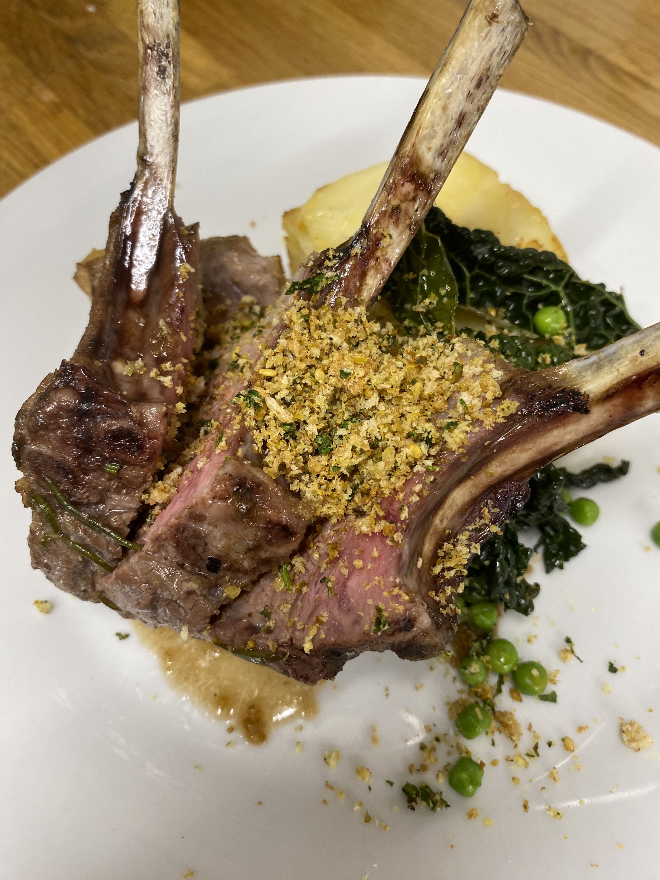 A white plate with cooked lamb chops garnished with herbs, served with peas, kale, and mashed potatoes.