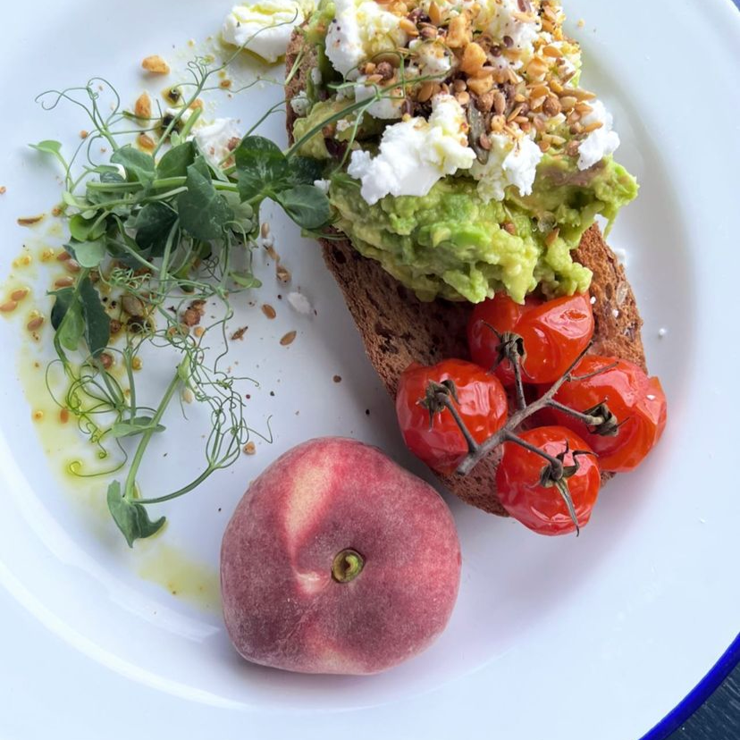 Open-faced avocado toast on a slice of whole grain bread, topped with cherry tomatoes, crumbled cheese, and chopped nuts, served with microgreens, a peach, and drizzled olive oil on a white plate.