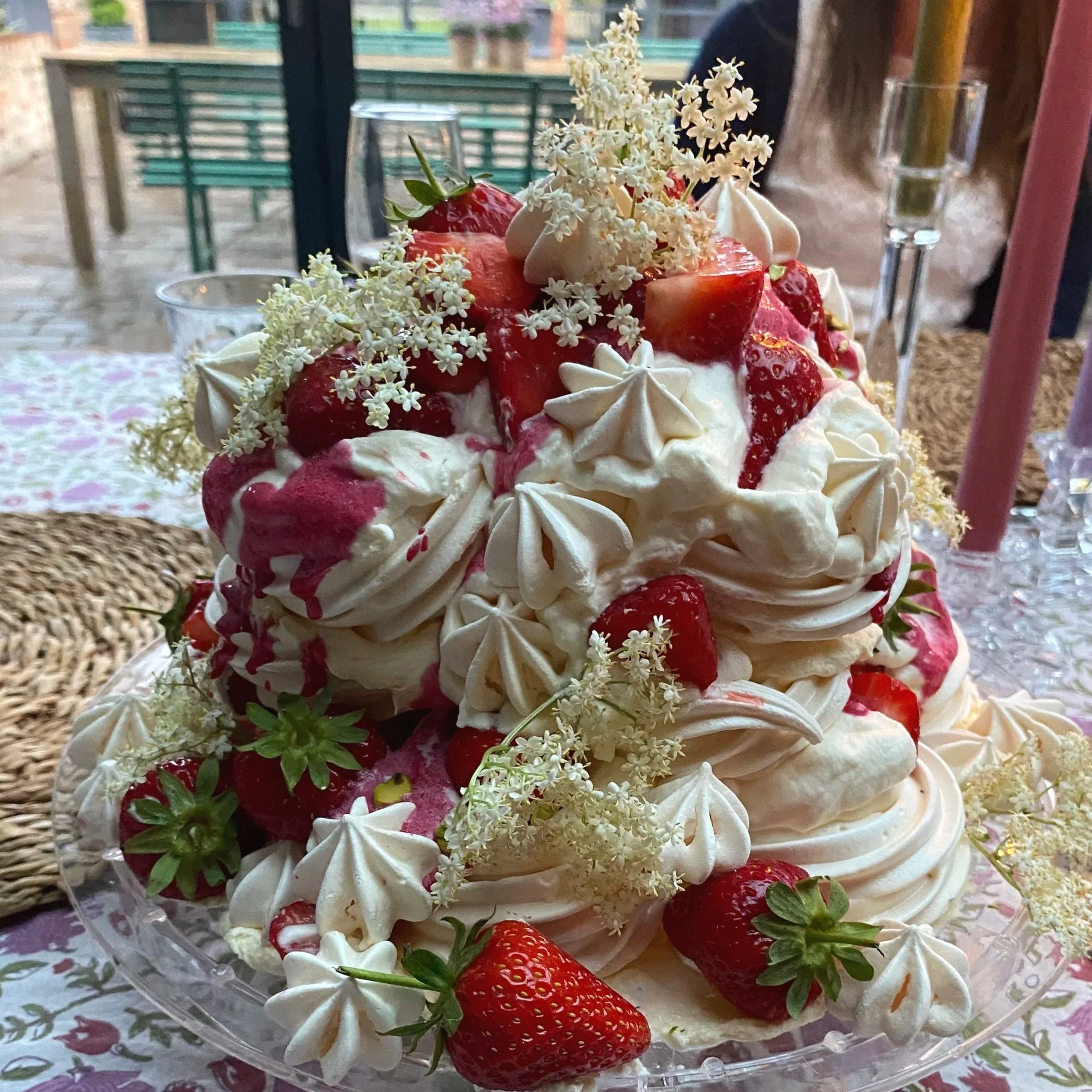 A layered cake decorated with strawberries, white flowers, and meringue kisses, with pink syrup drizzled on top.