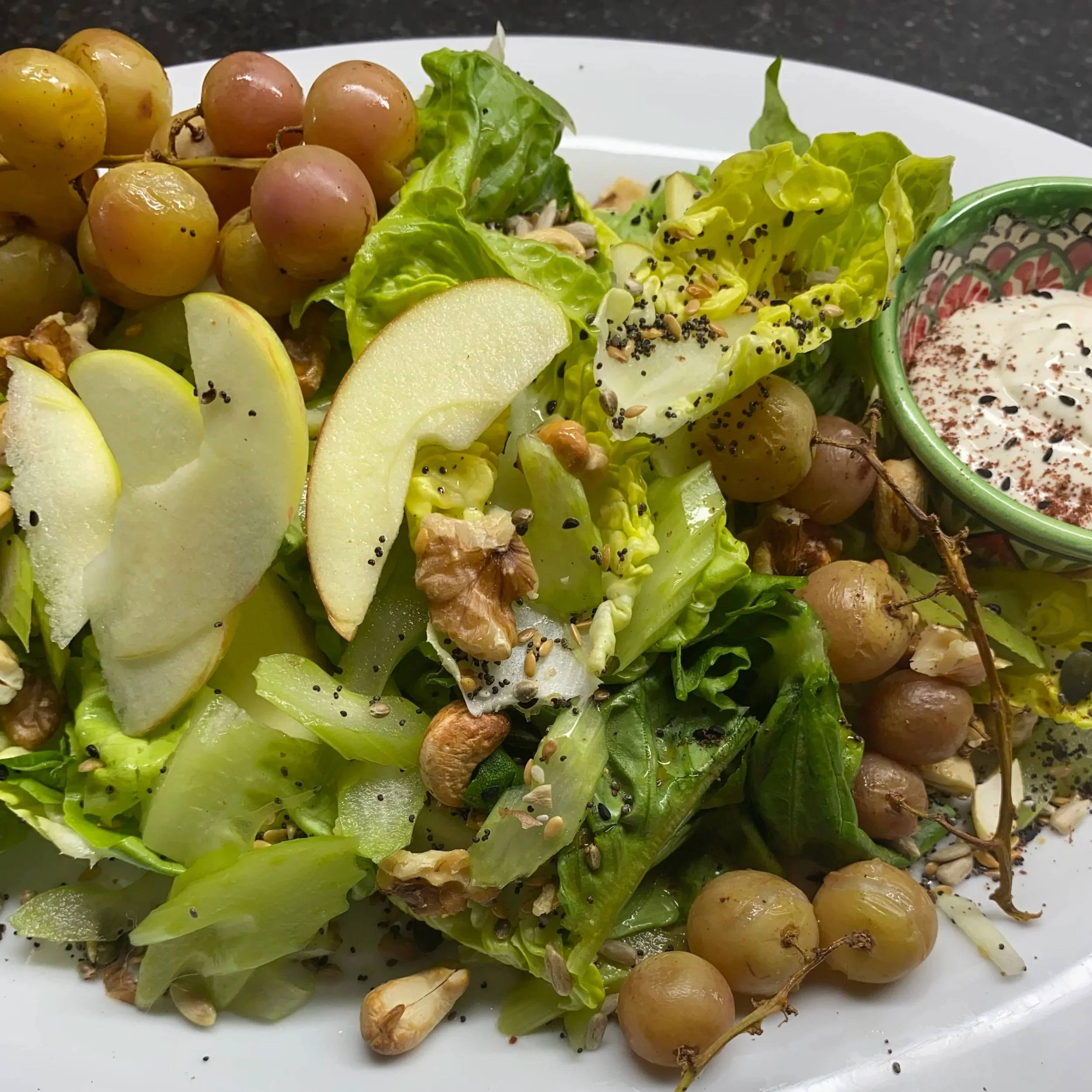 Mixed green salad with apple slices, grapes, walnuts, sunflower seeds, black sesame seeds, and a small bowl of salad dressing.