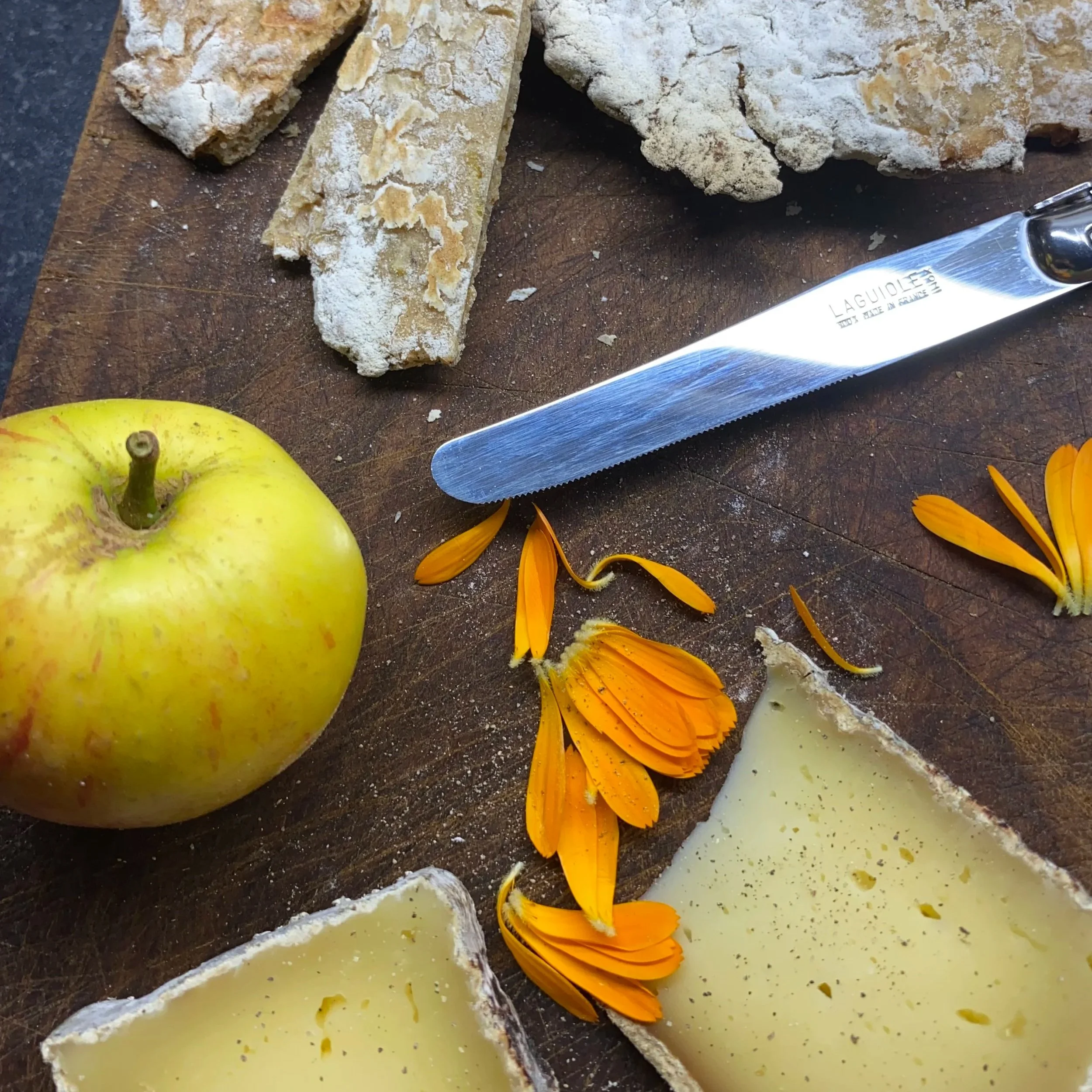 A wedge of cheese, an apple, an orange flower, a breadstick, and a bread loaf on a wooden cutting board with a cheese knife.