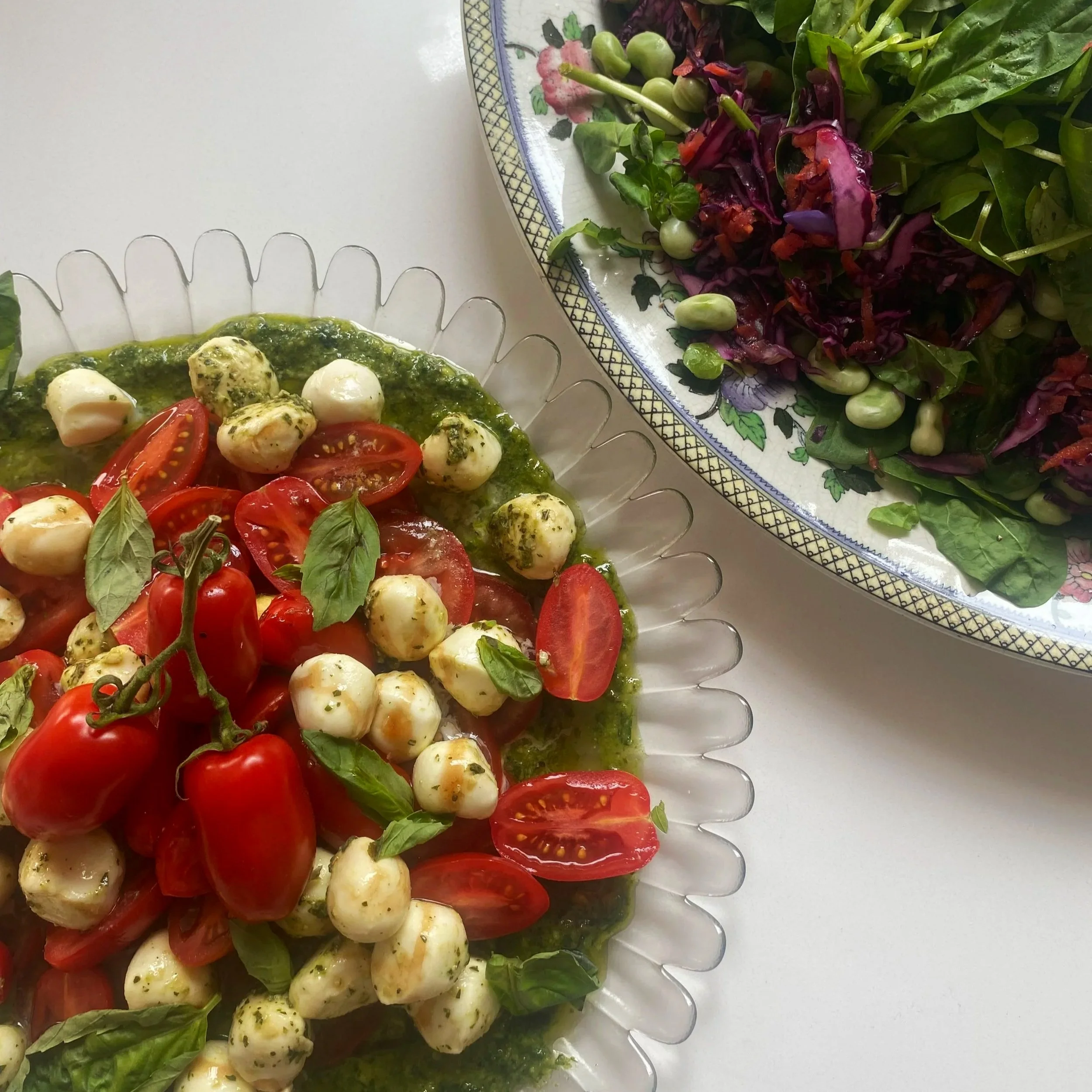 Two decorative plates of salad with cherry tomatoes, mozzarella balls, basil, and greens on a white surface.