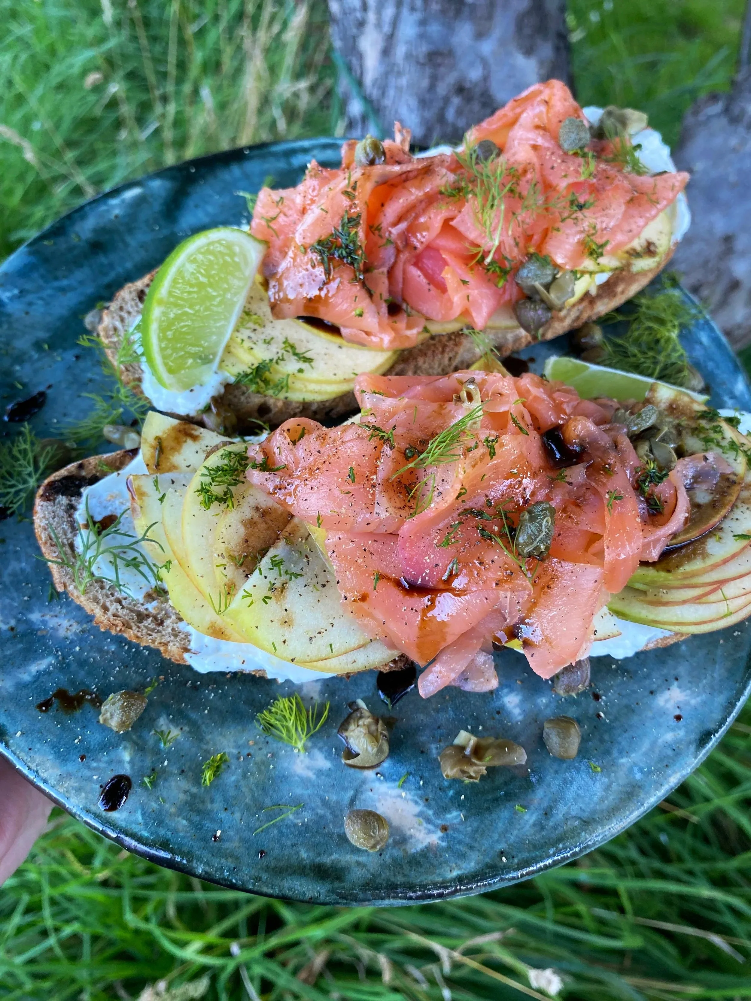 Two slices of bread topped with smoked salmon, sliced apples, capers, and dill, garnished with lemon wedges and herbs, served on a blue plate outdoors.