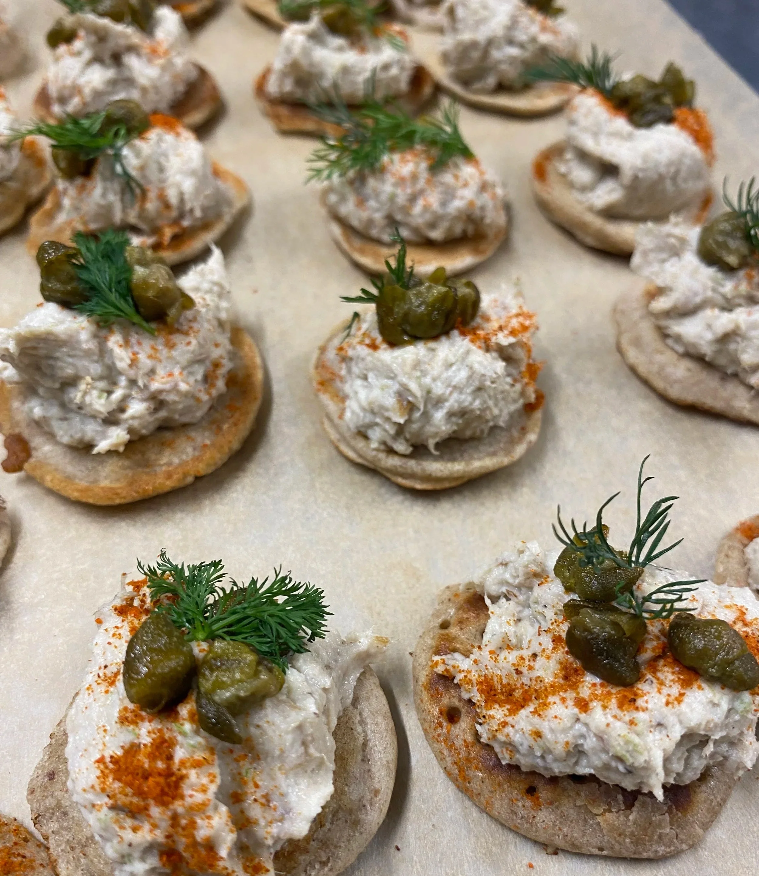 Close-up of several homemade crackers topped with a creamy chicken salad, garnished with pickles, fresh herbs, and paprika.
