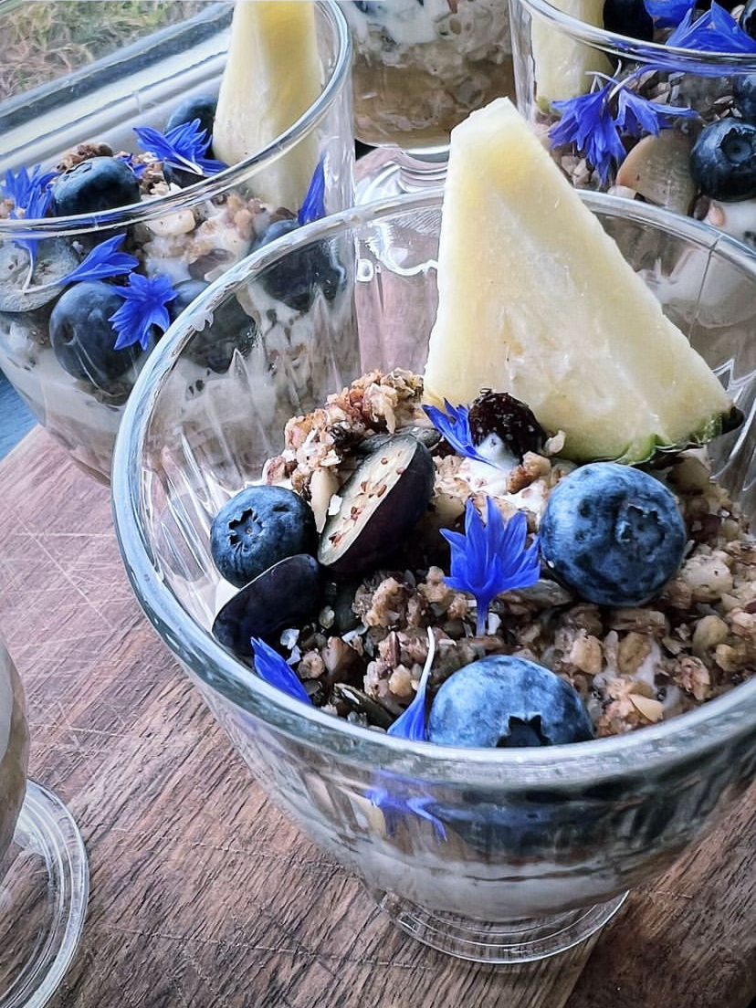 Glass bowl containing granola, blueberries, a wedge of melon, and blue edible flowers.