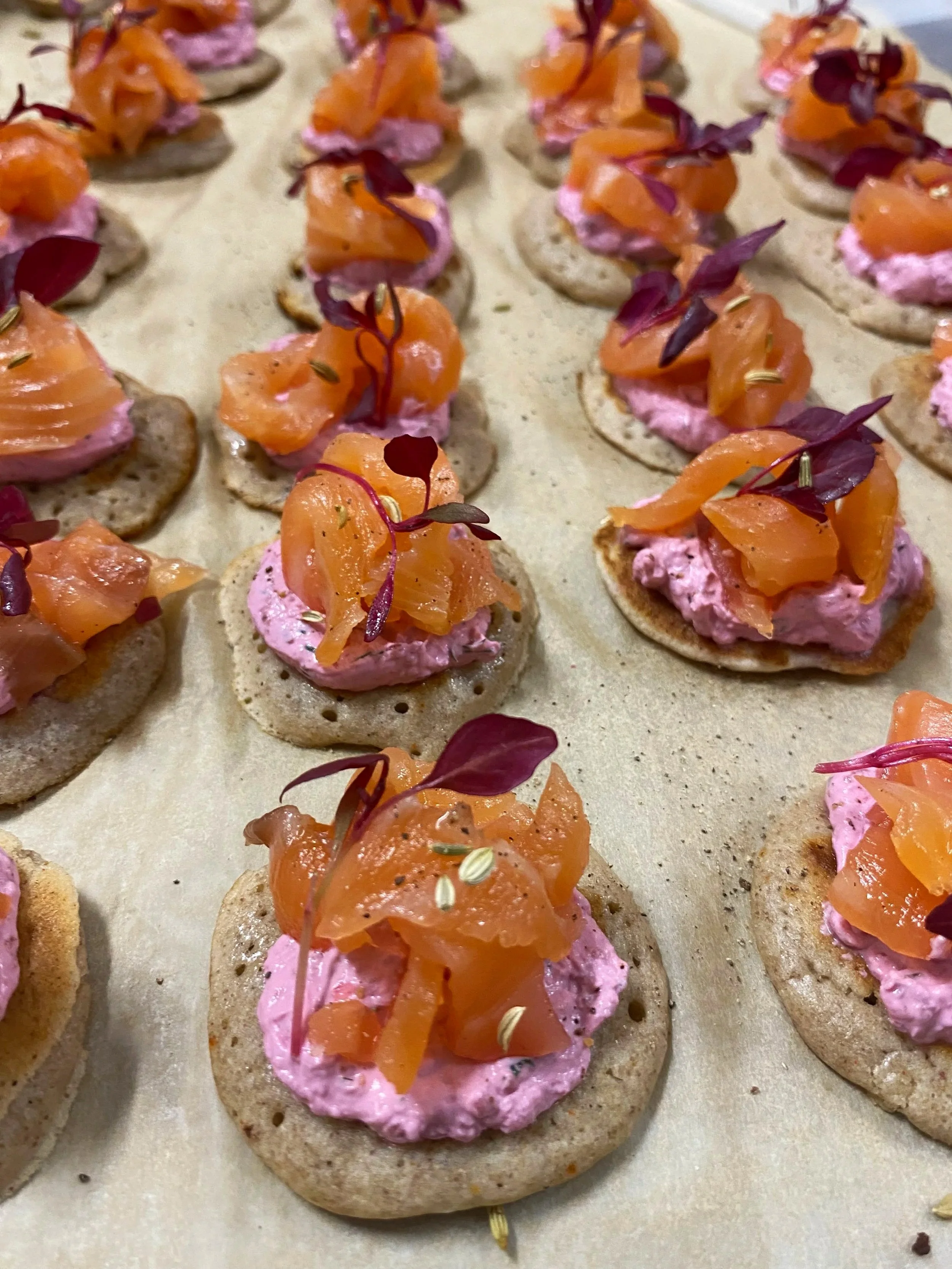 Close-up of canapés with pink cream, smoked salmon, microgreens, on round crispy crackers arranged on parchment paper.