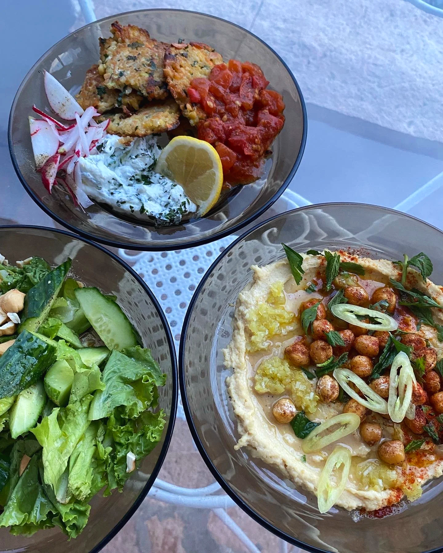 Three glass bowls of Mediterranean dishes on a table. One contains fresh salad with lettuce, cucumber, and nuts; another has hummus topped with chickpeas, lemon zest and paprika; the third has a vegetable stew with tomatoes and herbs. The bowls are o