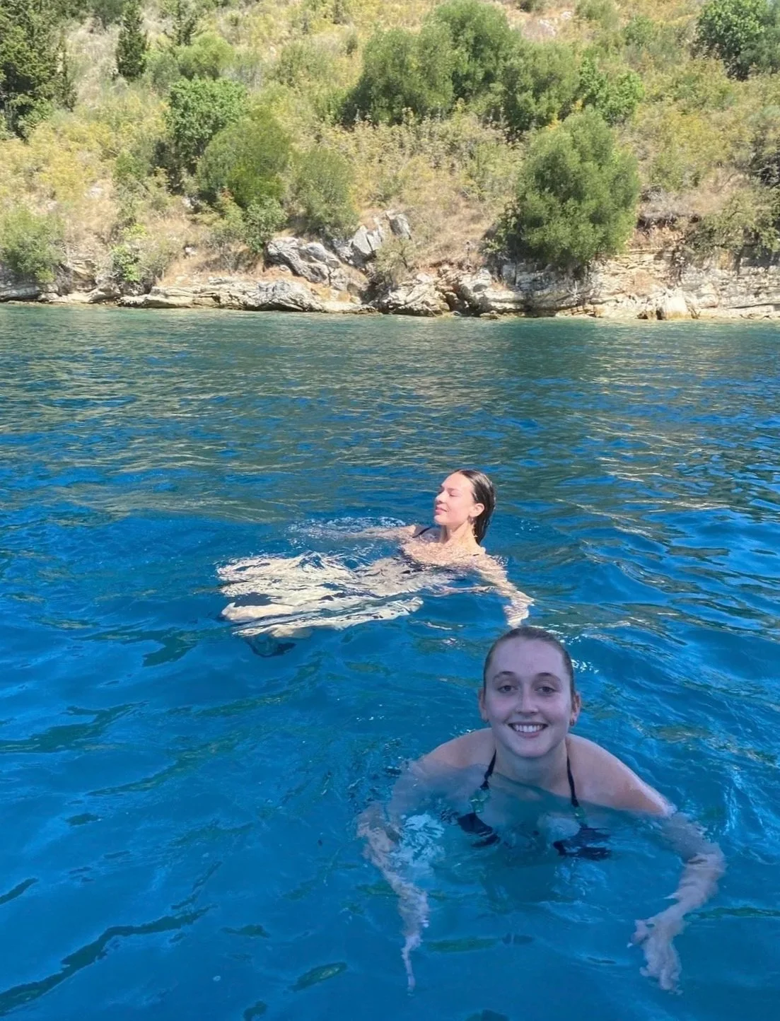 Two women swimming in a clear blue ocean in Corfu with green trees and rocky shoreline in the background.