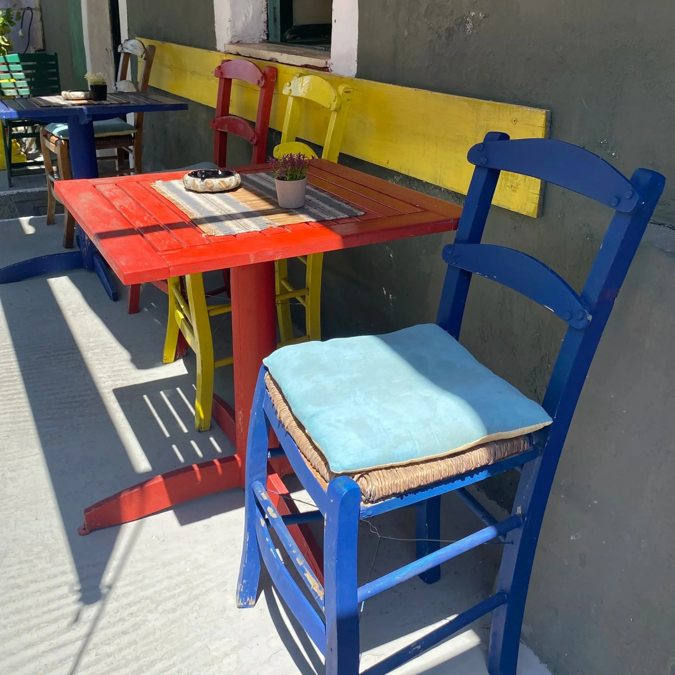 Colorful outdoor seating area with blue, red, and yellow wooden chairs and matching tables, a small potted plant, and a wall in the background