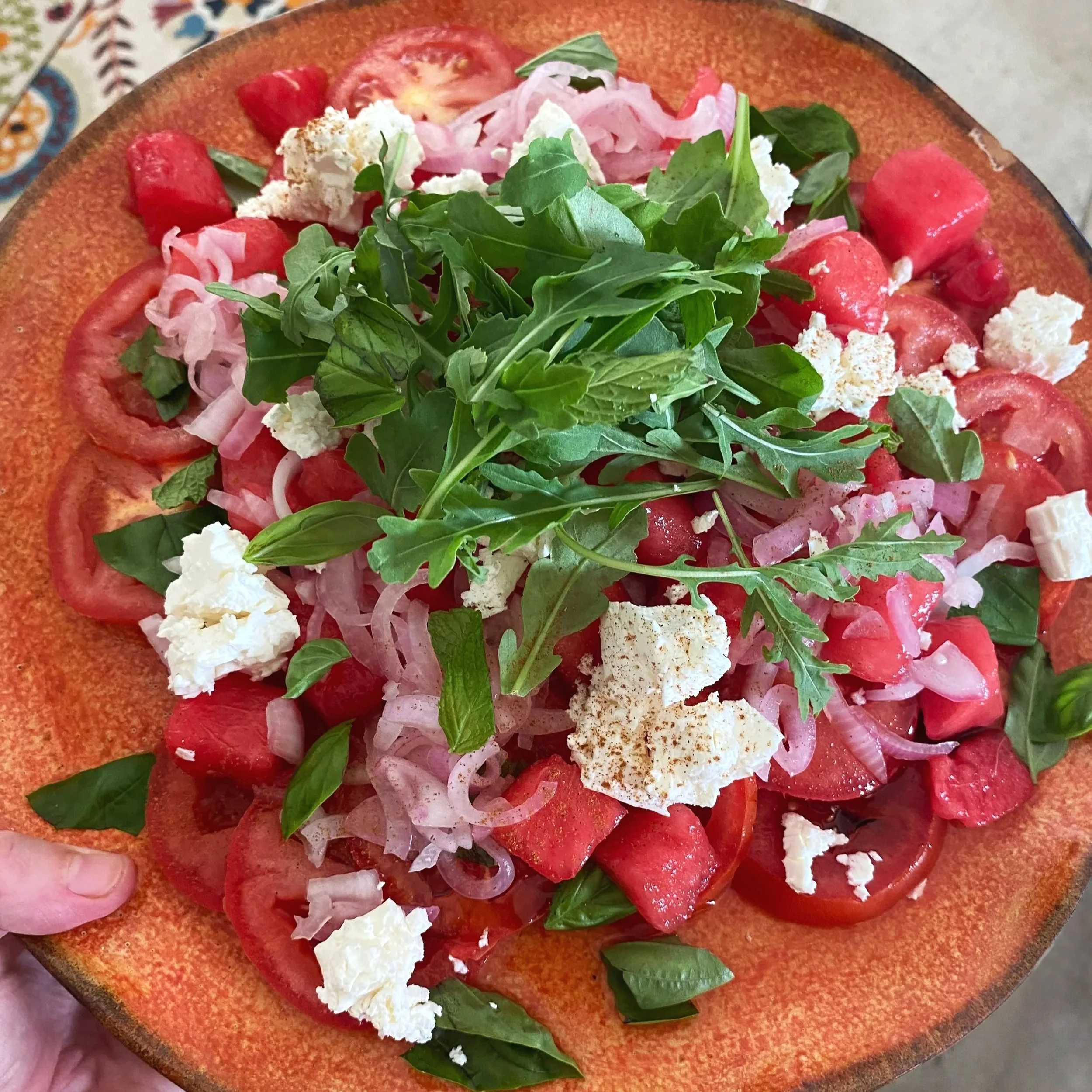 A salad with sliced tomatoes, sliced onions, crumbled white cheese, fresh green basil and arugula on an orange ceramic plate.