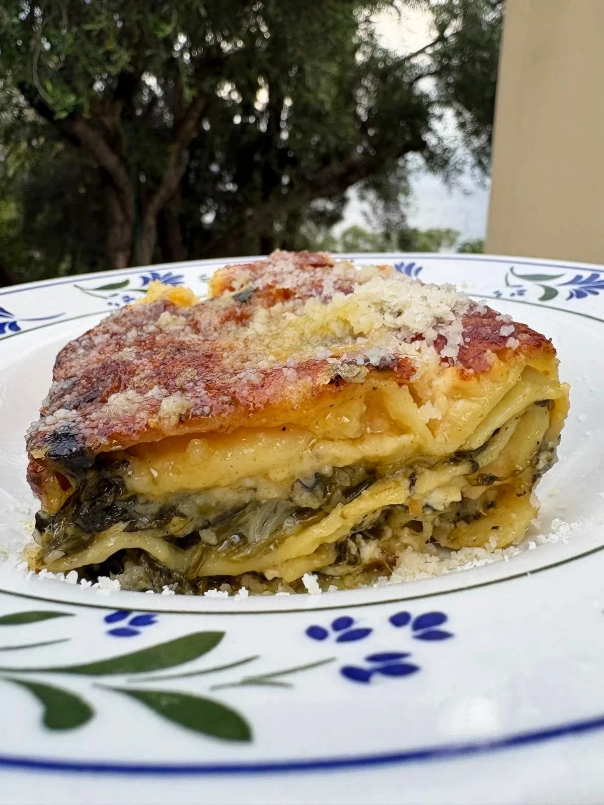 A slice of vegetable lasagna on a decorative white plate with blue and green floral patterns, set outdoors with trees in the background.