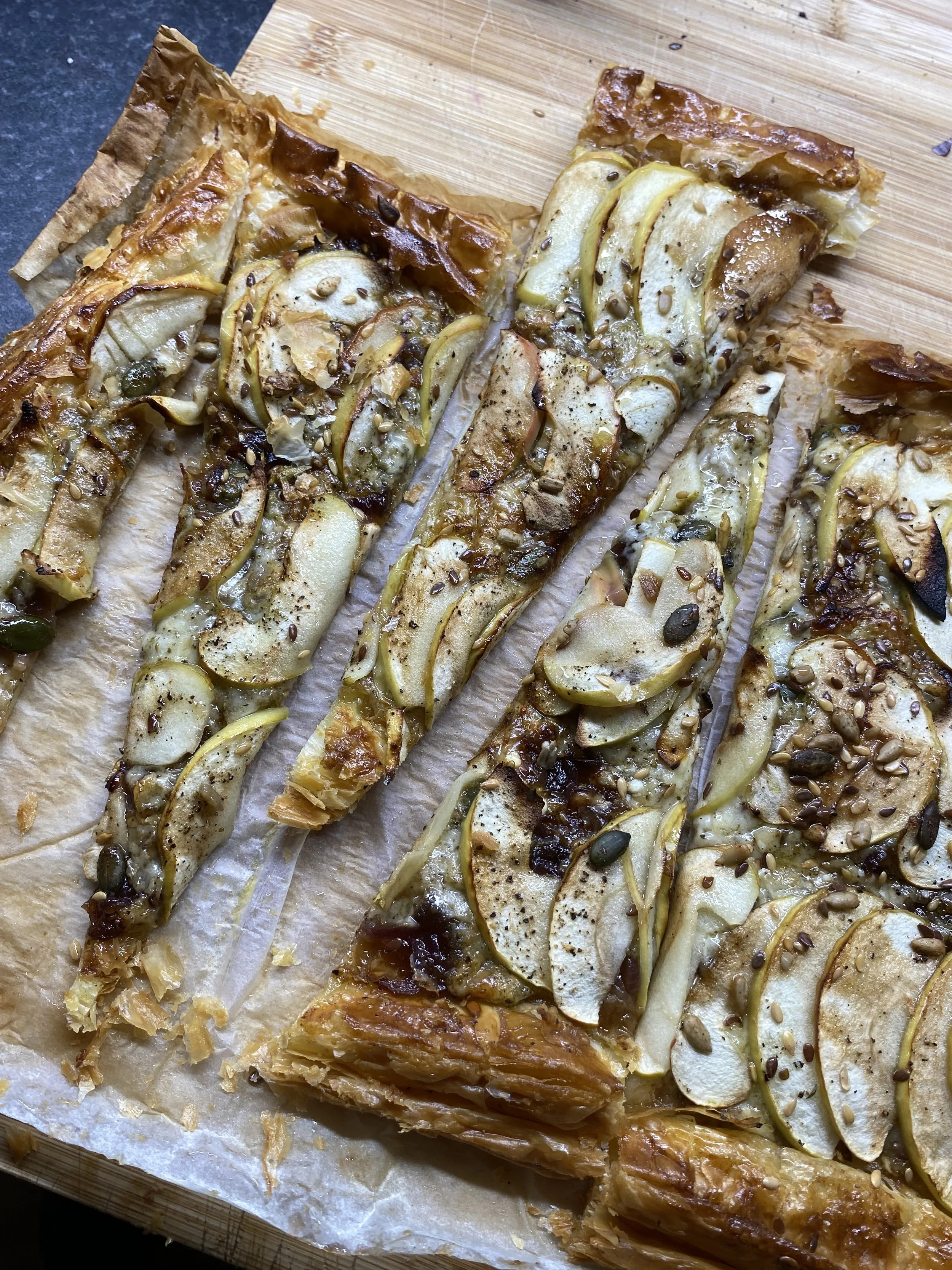 Puff pastry topped with sliced pears, seeds, and herbs on a wooden cutting board.