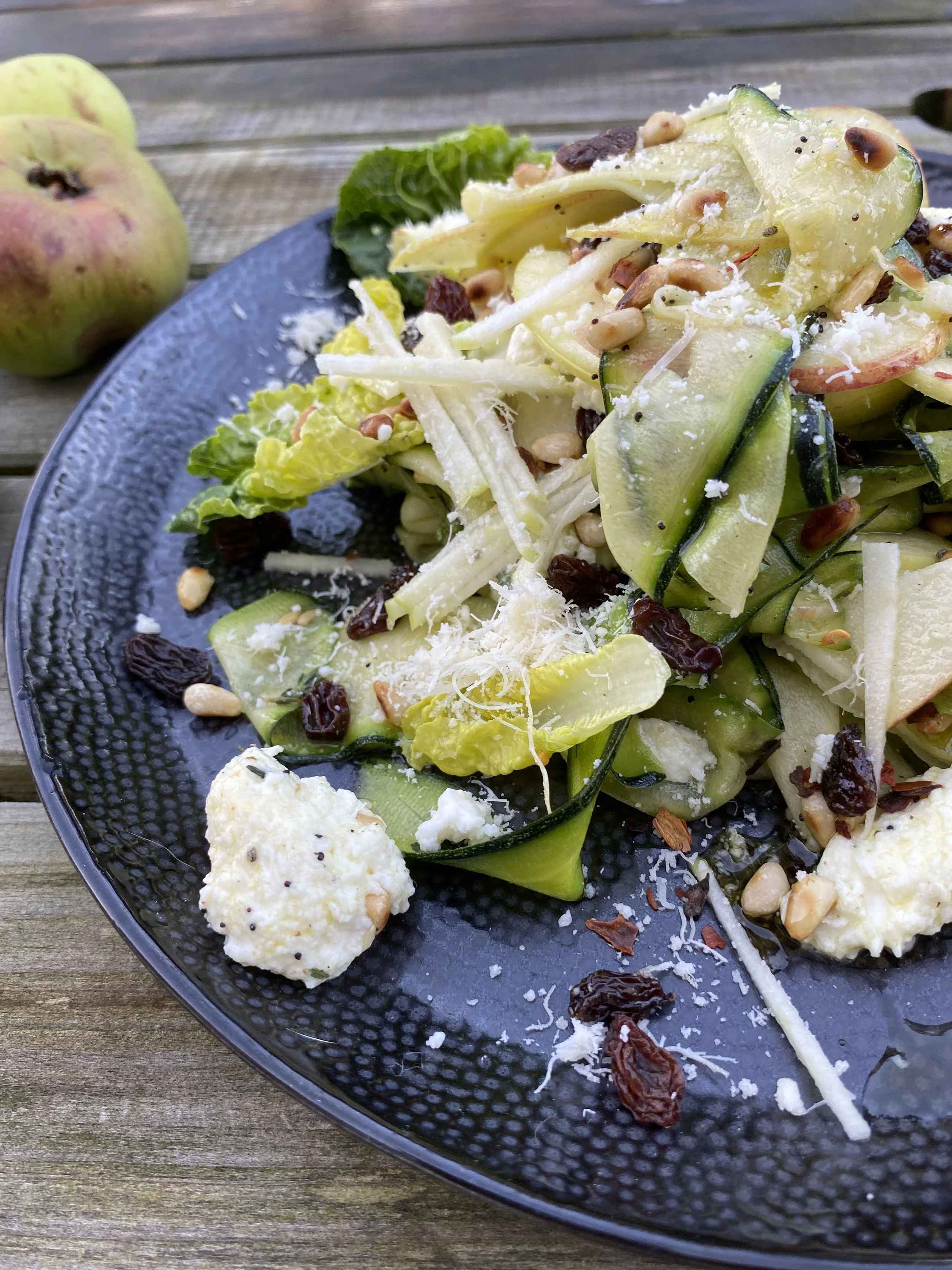 Cucumber and gem lettuce salad with sunflower seeds, dried cranberries, torn mozzarella and apple sticks on a black textured plate, placed on a wooden surface with an apple in the background.