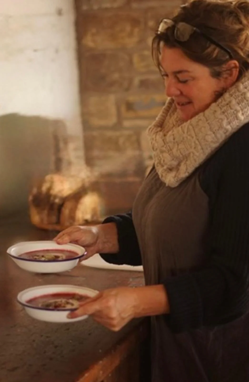 Woman with short hair and glasses on her head, wearing a tan scarf and black top, holding two bowls of soup in a cozy kitchen with brick walls.