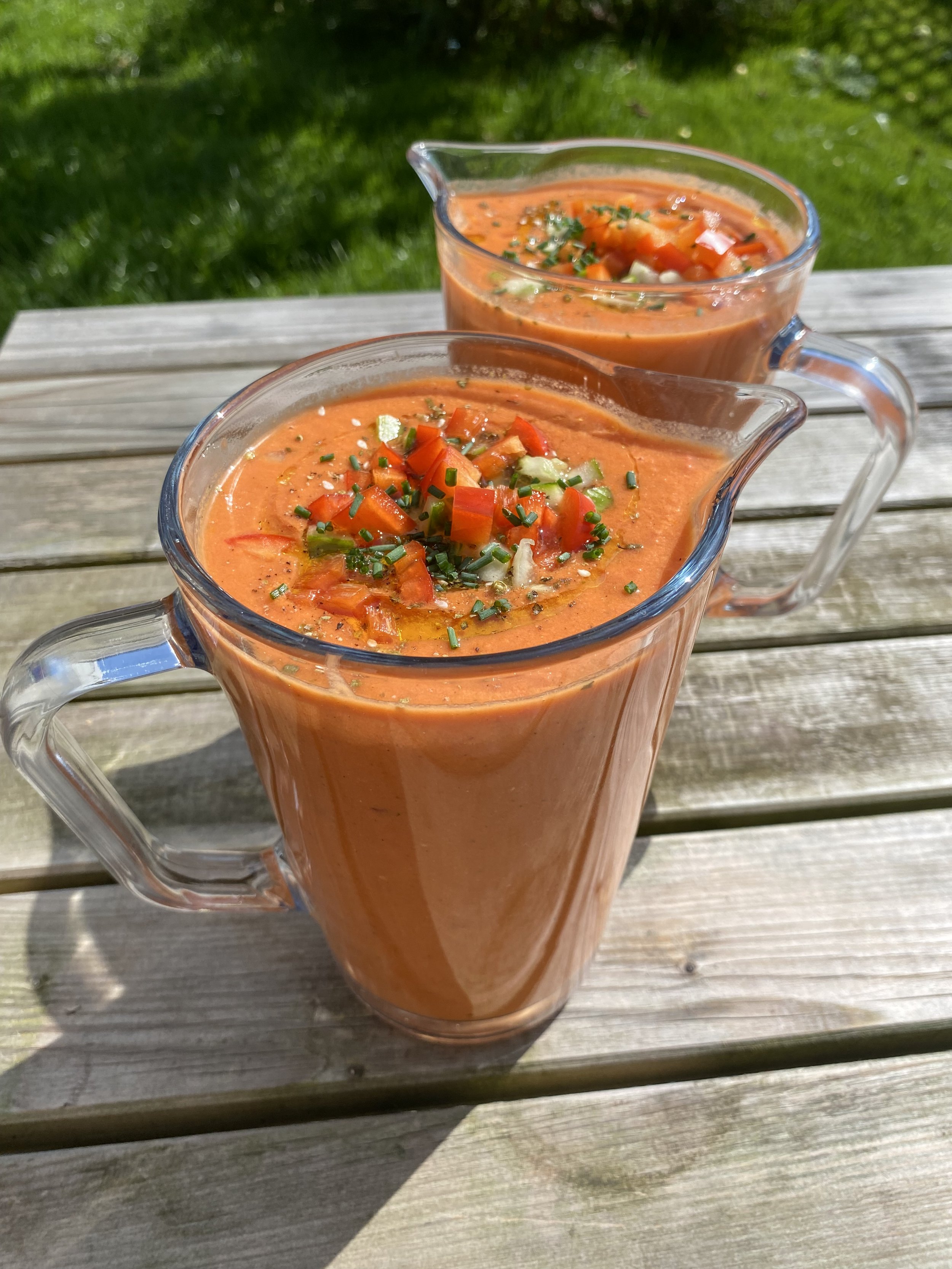 Two glass pitchers filled with tomato-based gazpacho garnished with chopped vegetables and herbs, placed on a wooden garden table with grass in the background.