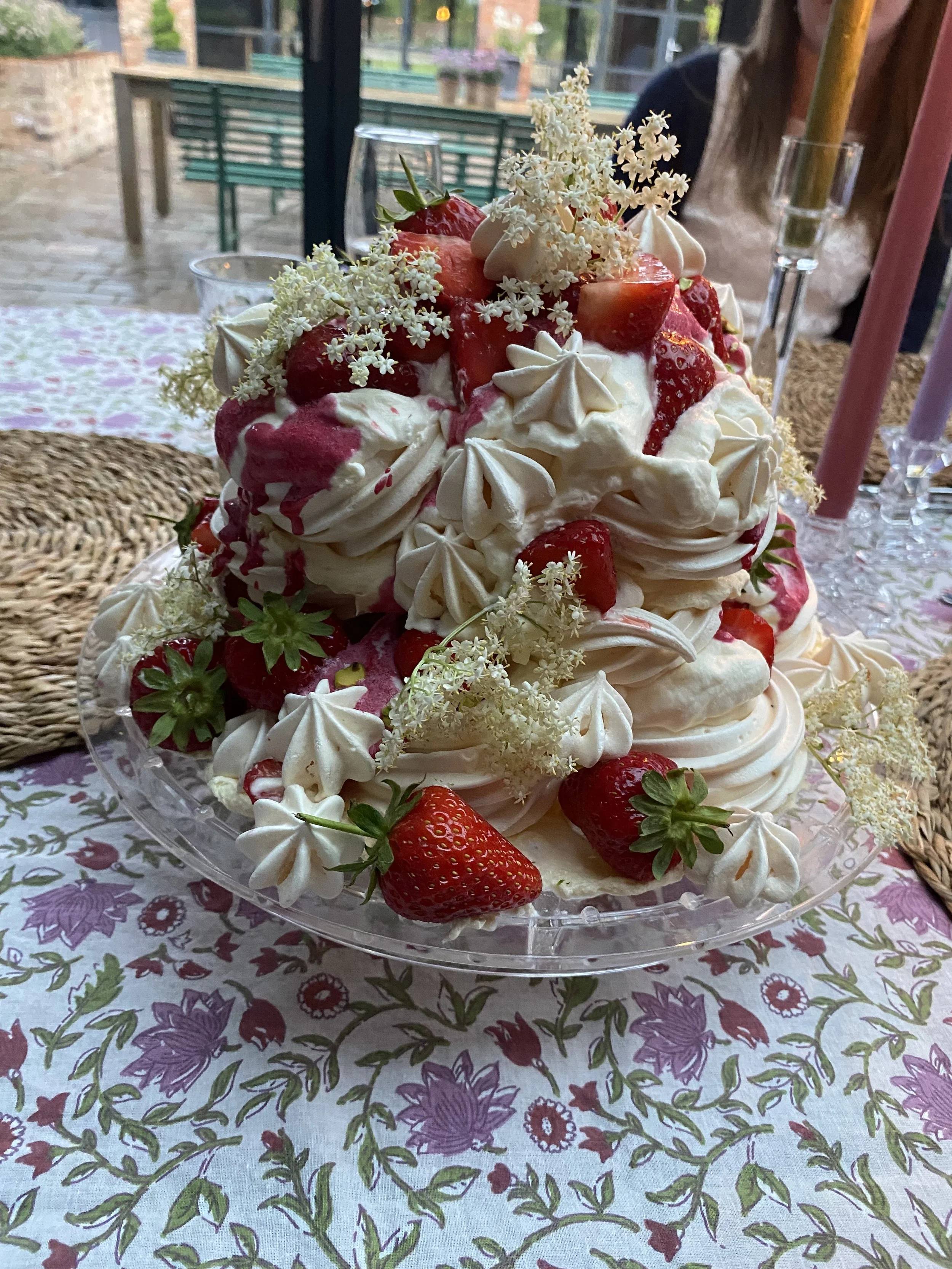 A layered strawberry pavlova cake decorated with strawberries, white flowers, and cream dollops on a glass platter, on a floral tablecloth.