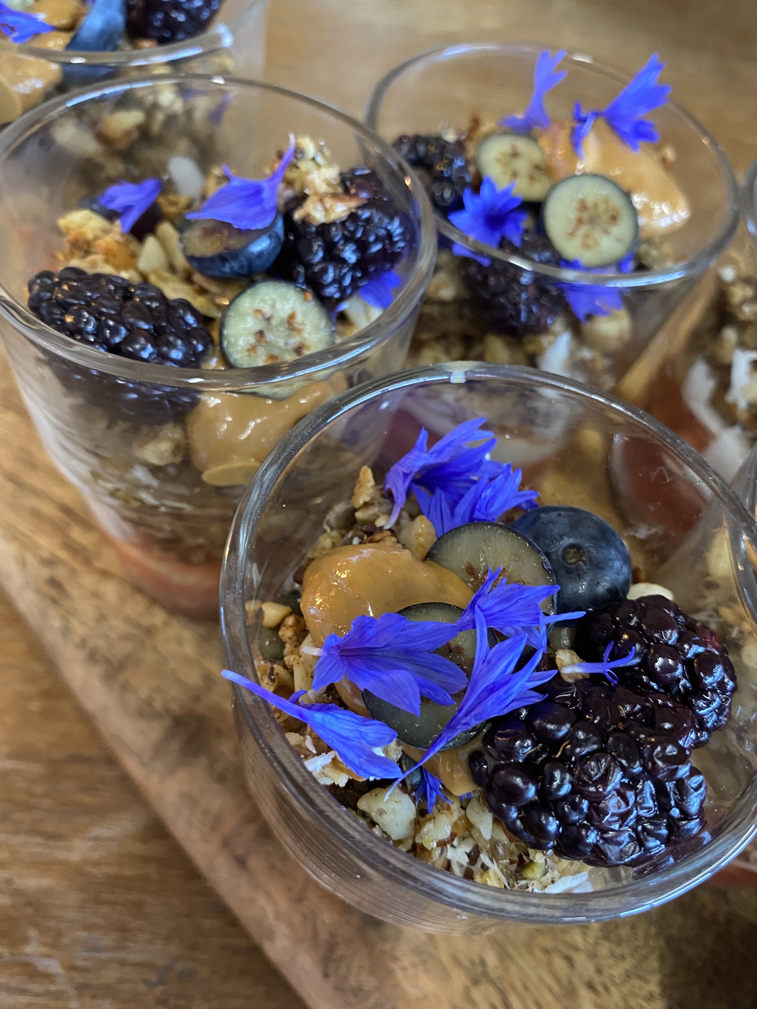 Dessert glasses filled with yogurt, blackberries, blueberries, sliced cucumbers, honey, granola, and edible blue flowers on a wooden surface.
