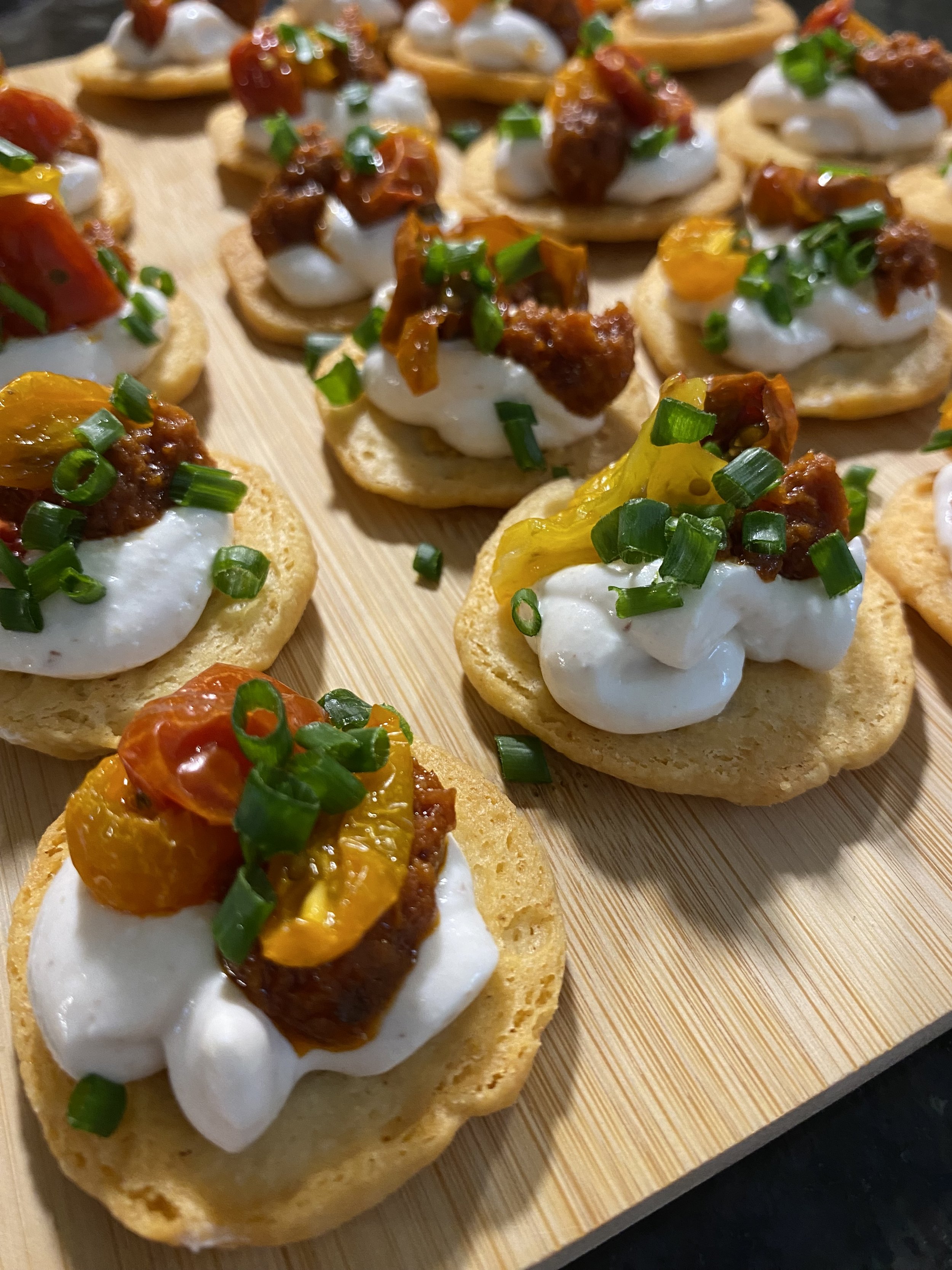 Close-up of several mini appetizers on a wooden board. Each consists of a toasted bread round topped with dollops of white cream, roasted cherry tomatoes, cooked meat pieces, and chopped green onions.