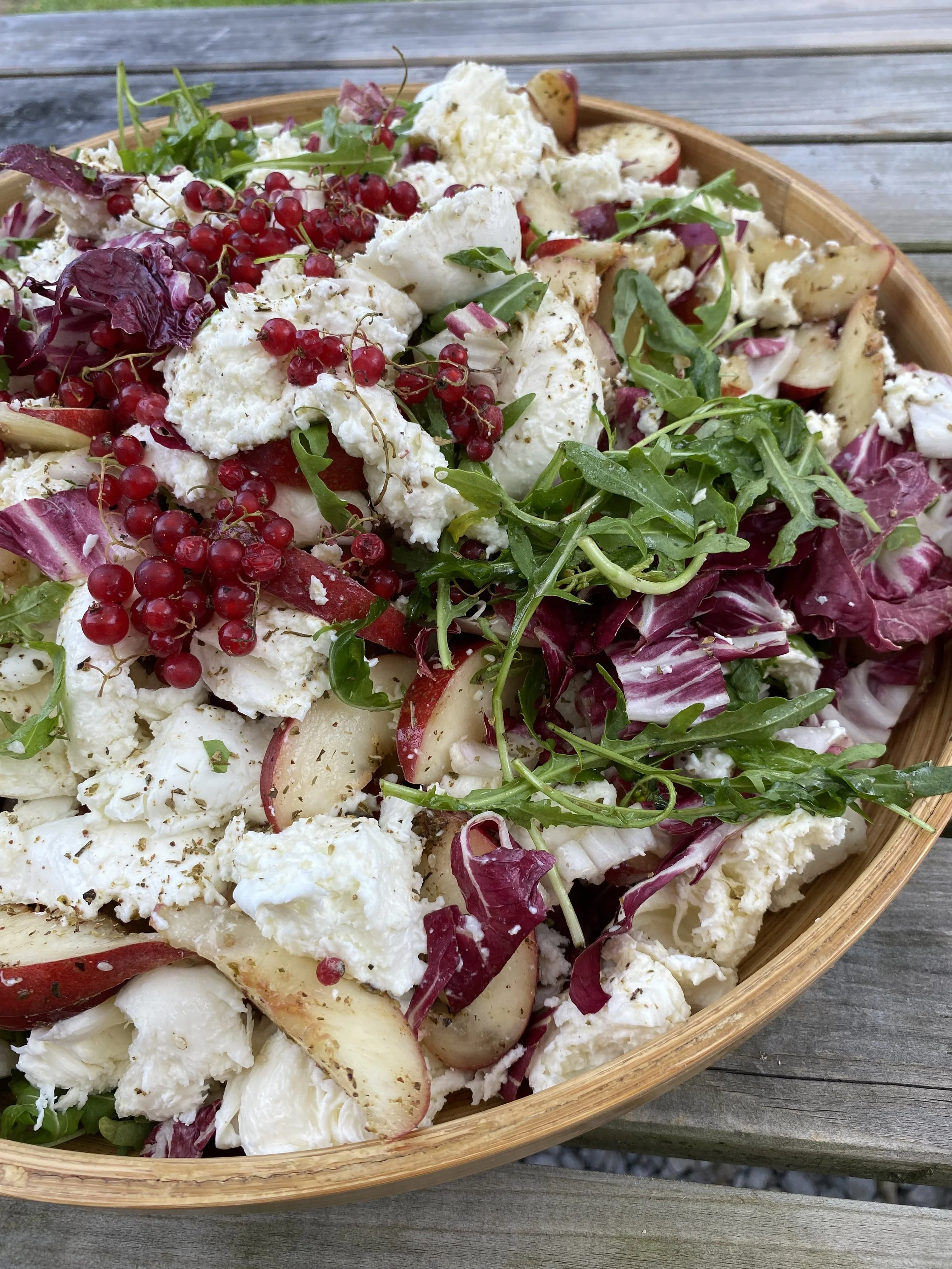 A wooden bowl filled with a fresh mixed salad containing rocket, radicchio, apple slices, crumbled feta cheese, and red currants on a wooden table.