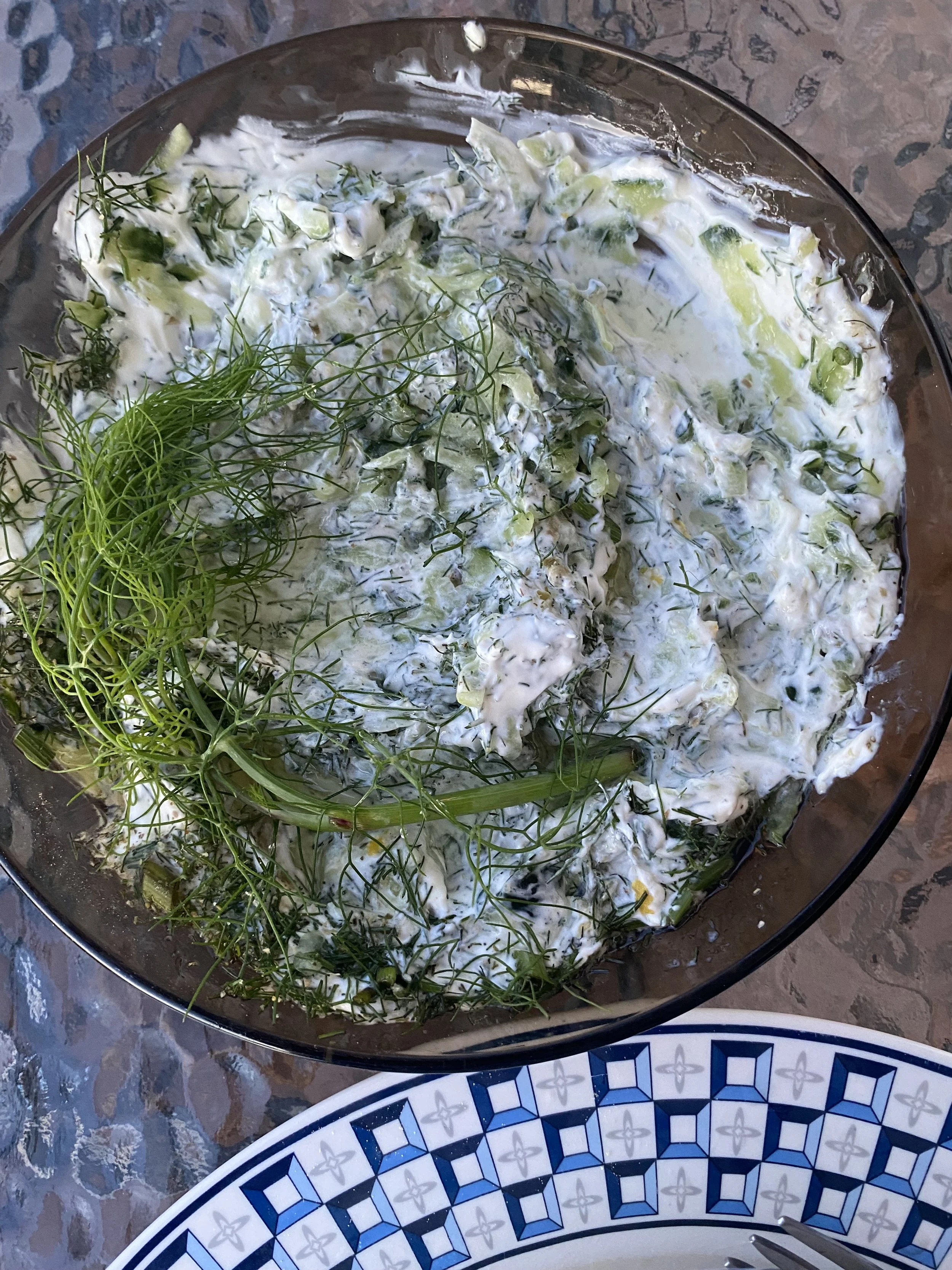 A bowl of creamy tzatziki garnished with fresh dill placed on a textured surface.