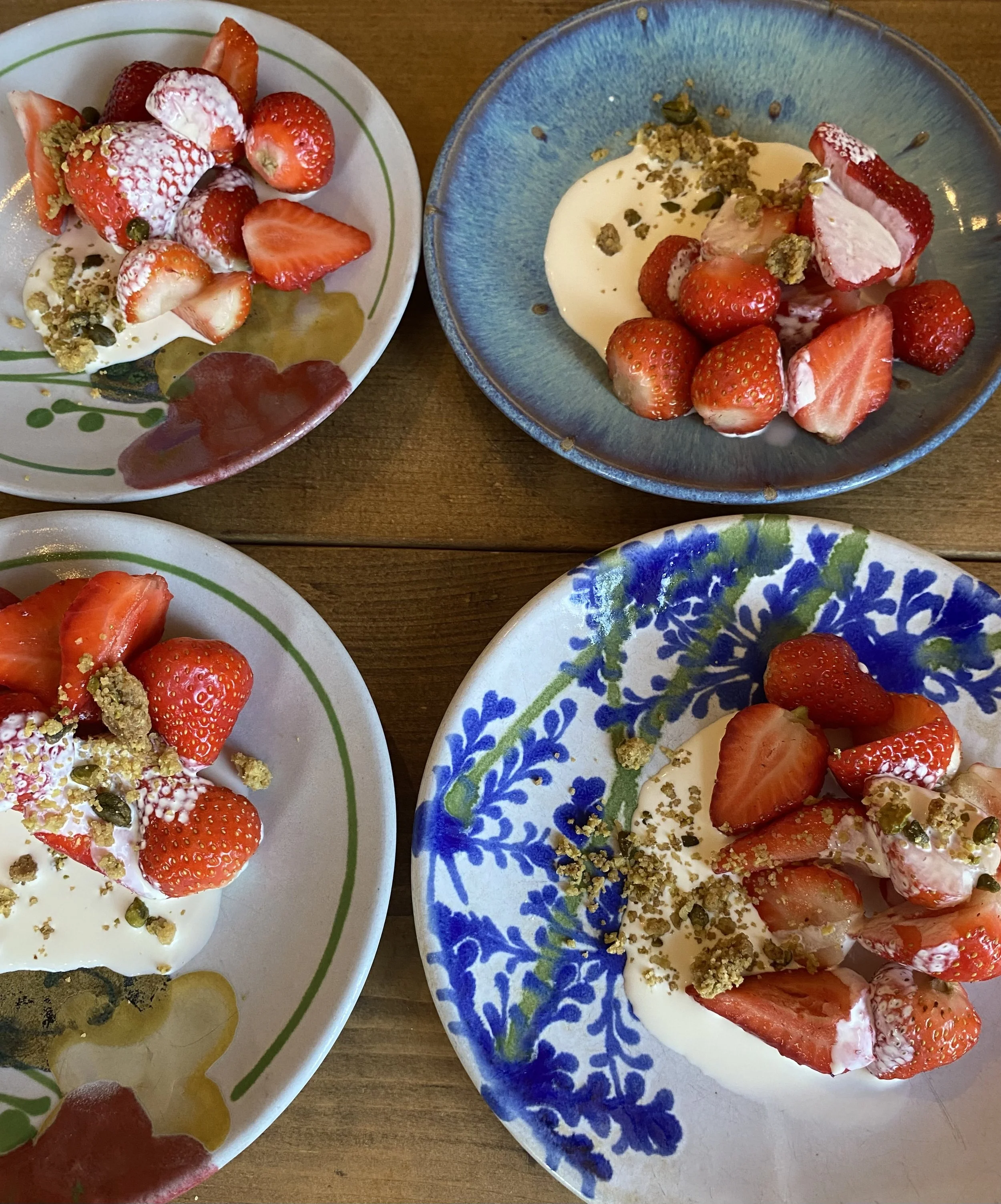 Bowls and plates of strawberry and cream dessert topped with crushed pistachios on a wooden table.