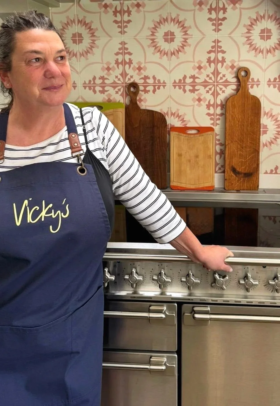 A woman wearing a striped shirt and an apron with 'Vicky's' written on it, standing in a kitchen next to a stove, with wooden cutting boards hanging on a patterned tile wall in the background.
